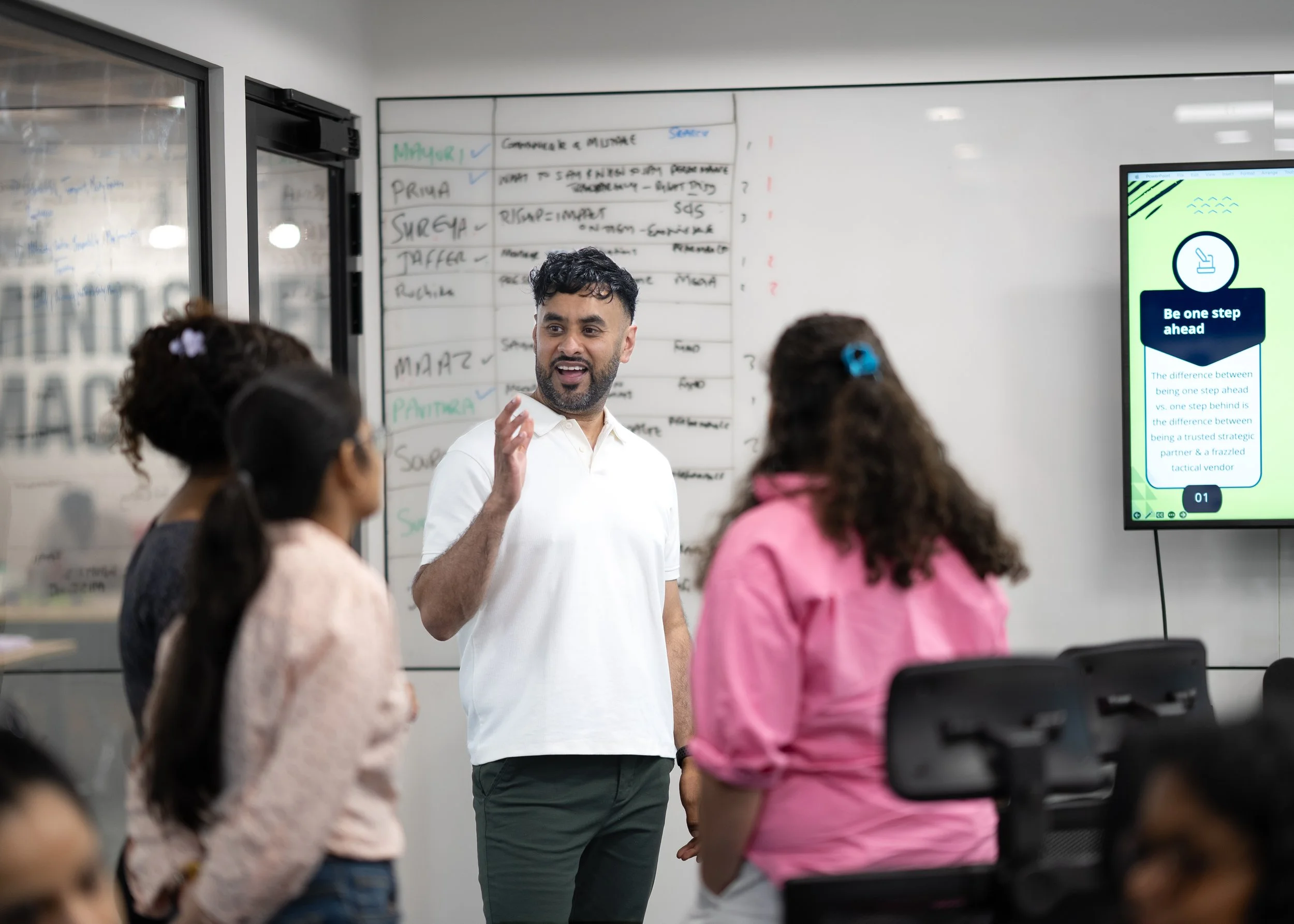A man in a white shirt is giving a presentation to a group of children in a classroom. There is a whiteboard with writing behind him and a large screen displaying a slide about being one step ahead.