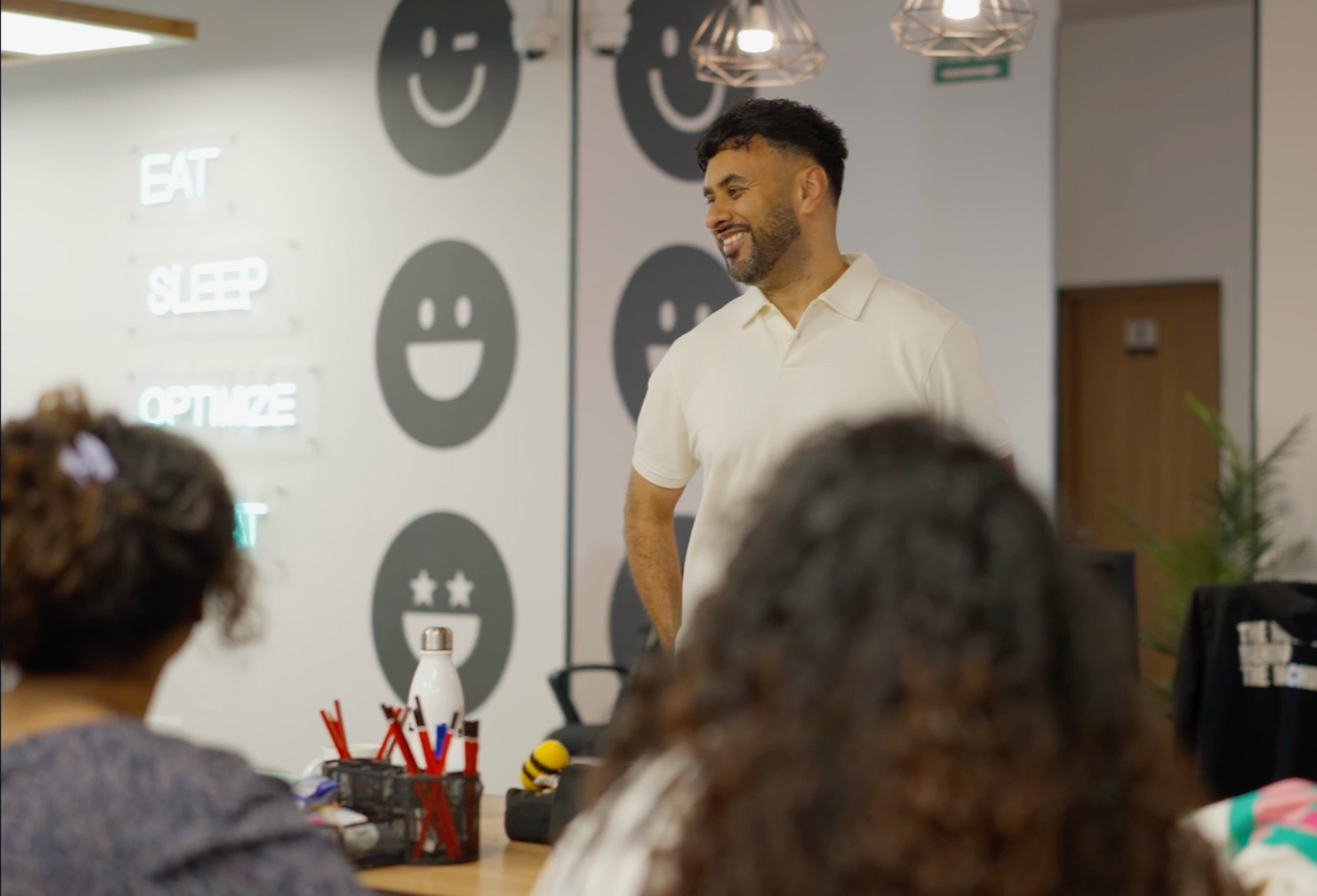 A man with dark hair and a beard smiling while standing in front of a wall with smiley face symbols and words like 'EAT', 'SLEEP', and 'OPTIMIZE'. The man is wearing a white polo shirt and appears to be speaking or presenting to a group of people sitting at a table.