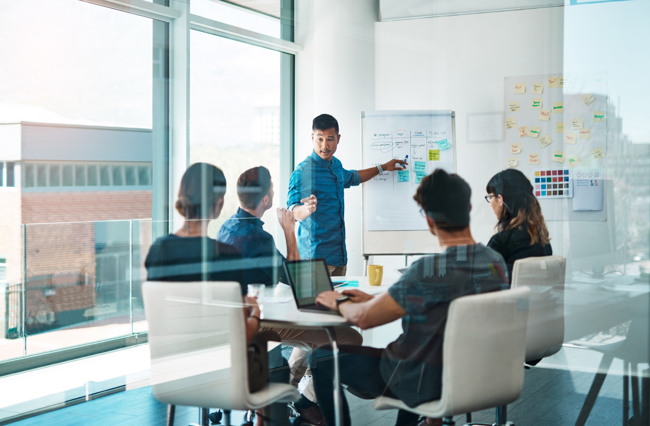 A man presents in front of a group of five people in a modern office with large windows. The presenter points at a flip chart, and the attendees are engaged, some with laptops open. The wall has colorful sticky notes and charts.
