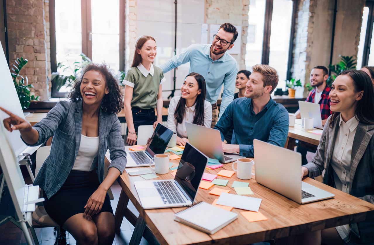 A diverse group of coworkers are in a modern office, smiling and engaging in a collaborative discussion around a table covered with laptops, notebooks, sticky notes, and mugs, with large windows and brick walls in the background.