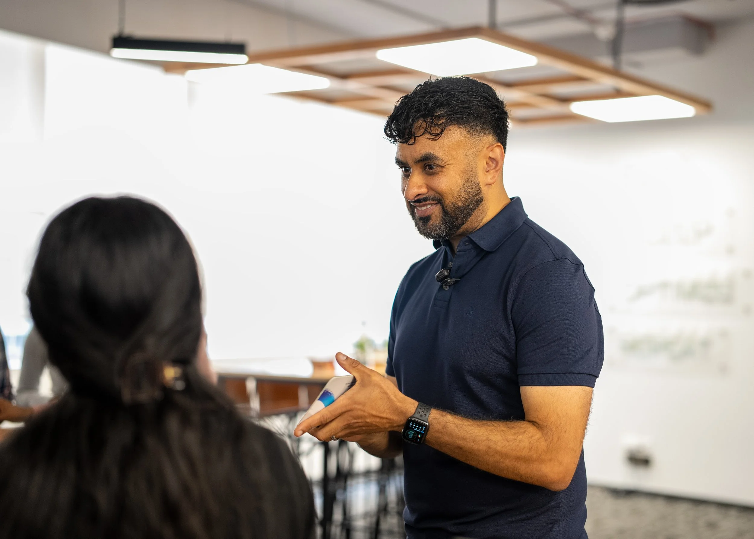 A man with dark, curly hair and a beard wearing a navy blue polo shirt, smiling and talking to a person with long dark hair in an indoor setting with bright lighting and modern ceiling fixtures.
