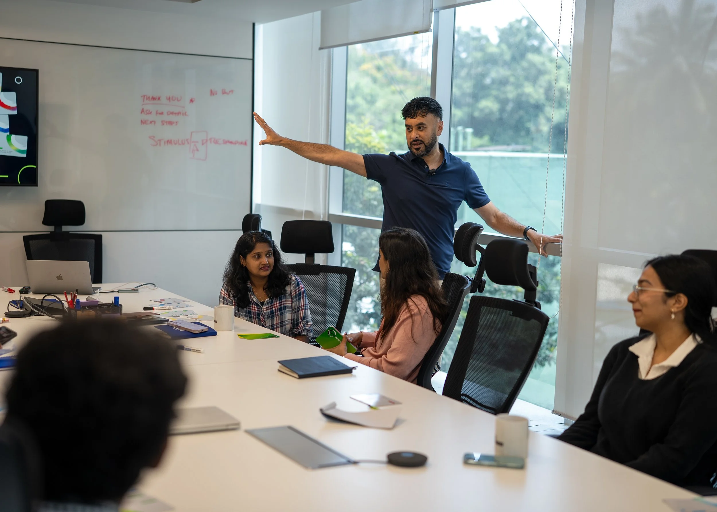 A group of diverse people sitting and listening in a meeting room with large windows, a man standing and gesturing to the whiteboard with red writing, and various notebooks, laptops, and cups on the table.