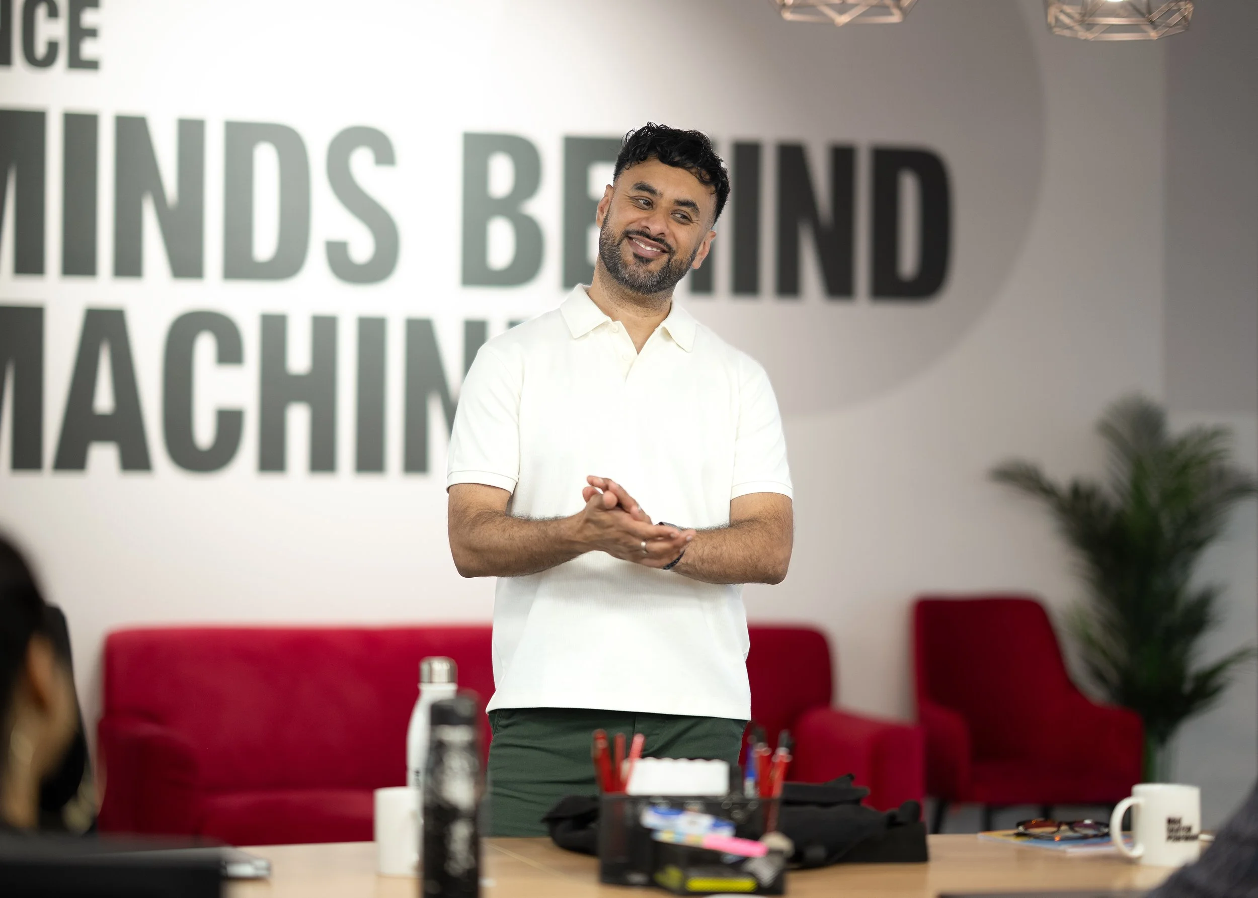 A man with dark hair and a beard, wearing a white polo shirt, standing and smiling in a conference room with a large sign that reads 'LIND'S BEYOND ACHIEVEMENT' behind him. There are red chairs, a table with various items, and some greenery in the background.