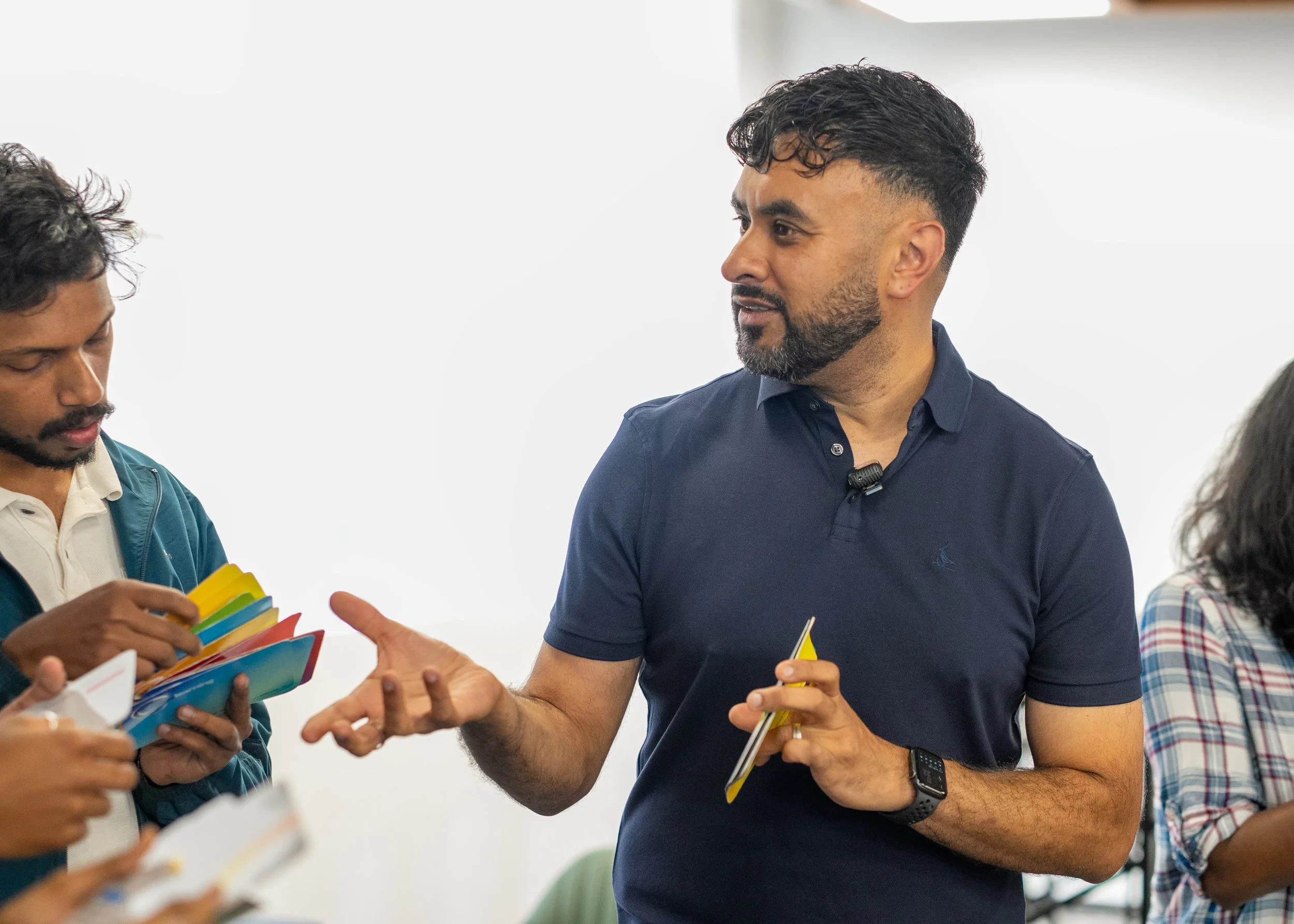 A man with dark hair, beard, and a blue polo shirt speaking to a group of people at a meeting. The woman on the right is holding a notebook, and another person on the left is holding a colorful fan of paper sheets. The background is plain white with soft lighting.