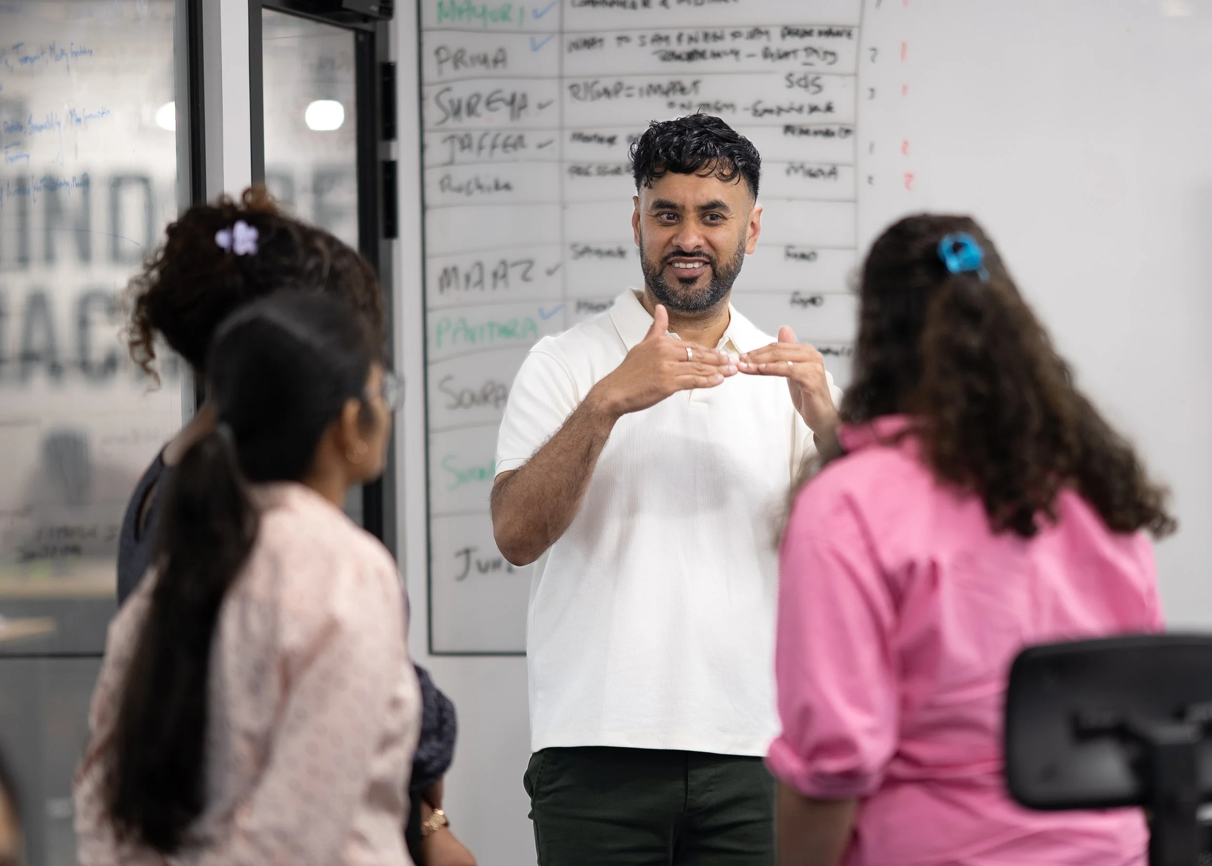A man speaking to a group of young children in a classroom, standing in front of a whiteboard with handwritten notes and schedules.