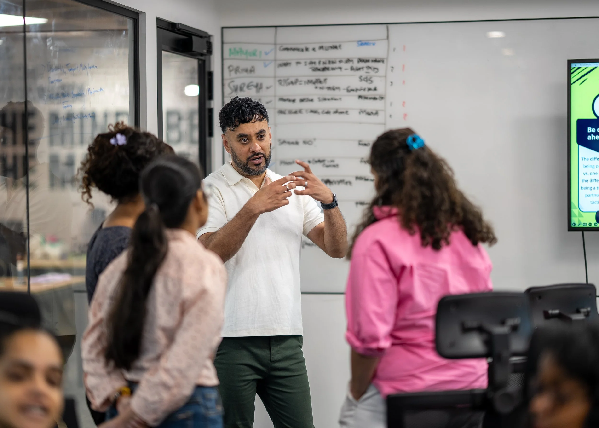 A man in a white shirt is standing and speaking to a group of children in a classroom. The children are listening attentively, some with hair accessories. There is a whiteboard with writing behind the man and a monitor displaying a colorful presentation to the right.