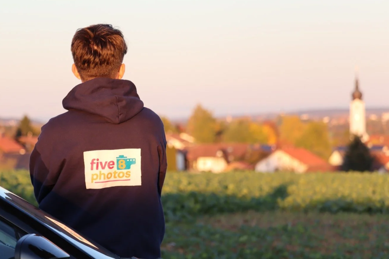 Junge mit hoodie, auf dem 'five 8 photos' steht, blickt auf eine ländliche Landschaft bei Sonnenuntergang