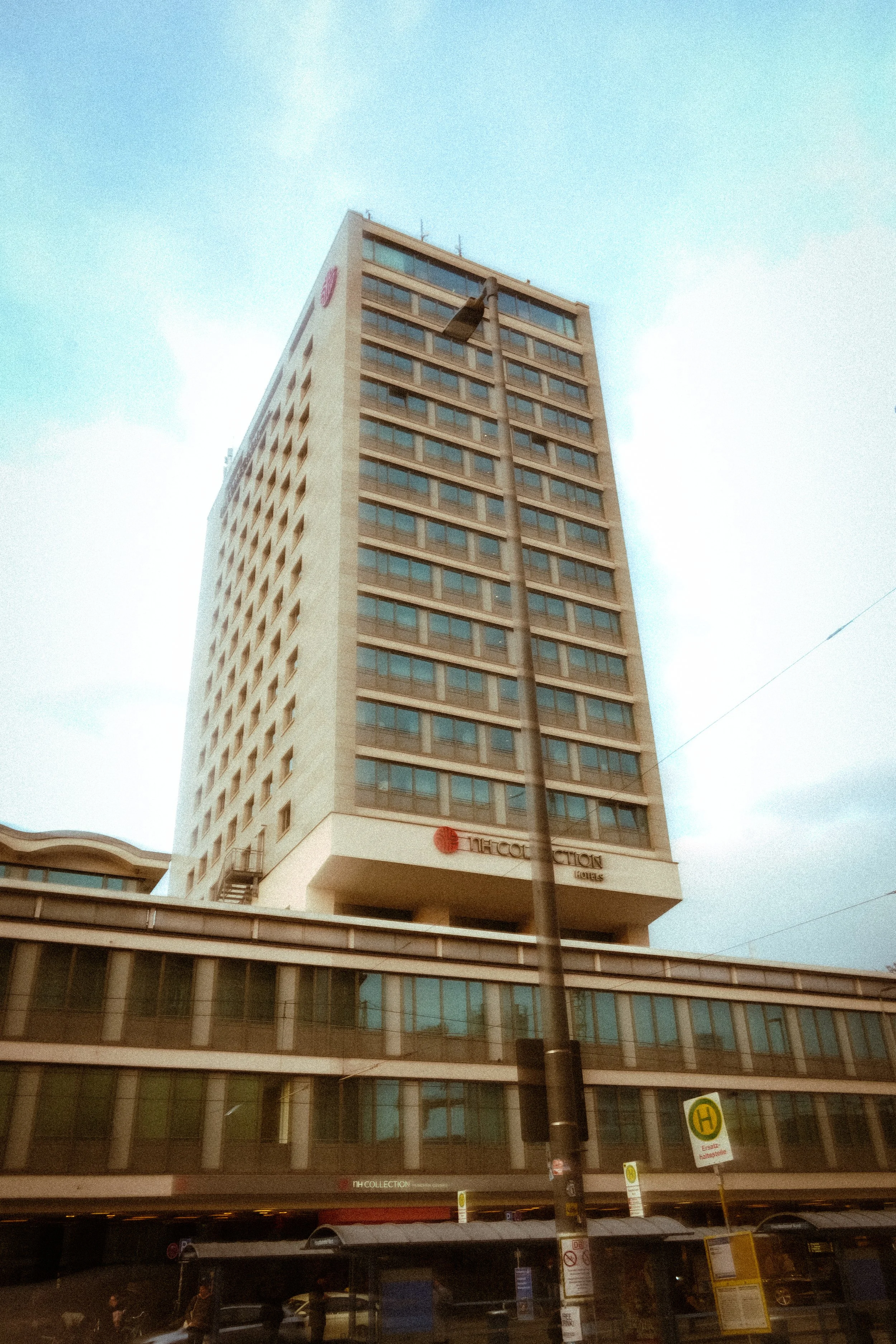 Mehrstöckiges Hotelgebäude mit großen Fenstern, ein Laternenpfahl davor und eine Bushaltestelle mit Linienbussen und Wartehäuschen. Es gibt auch Fahrsportrampen und Werbeschilder an der Bushaltestelle.
