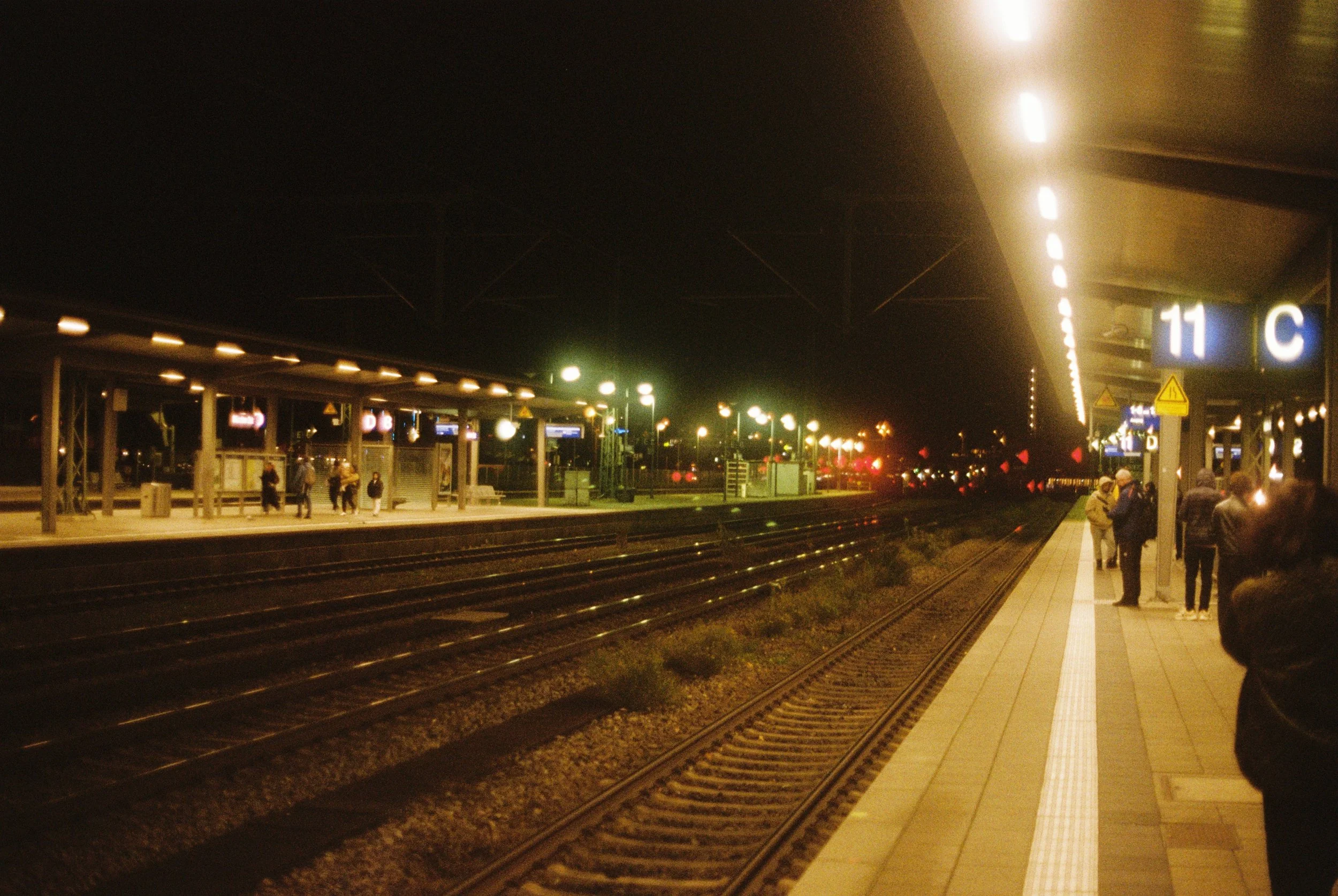 Nachtaufnahme eines Bahnsteigs mit Wartenden, Beleuchtung und Gleisen, am rechten Bildrand eine Gruppe von Personen, im Hintergrund Hinweise auf Züge und Signale.