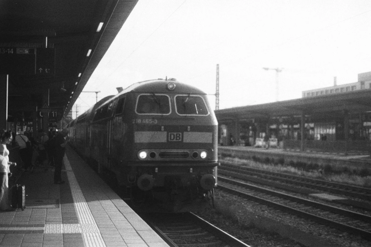 Ein schwarzer und weißer Zug steht am Bahnhof, mit Passagieren auf dem Bahnsteig, im Hintergrund sind Baukräne und ein Gebäude sichtbar.