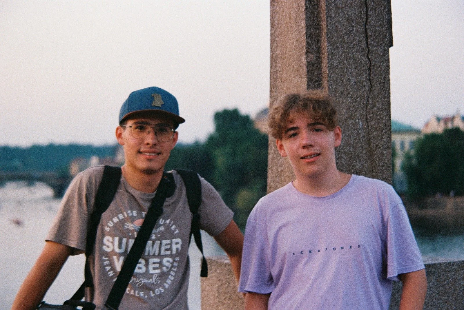 Zwei junge Männer stehen auf einer Brücke am Fluss bei Sonnenuntergang. Eines trägt eine Latzhose mit Krawattenmuster und ein Basecap, das andere ein lila T-Shirt. Im Hintergrund sind Bäume und Gebäude sichtbar.
