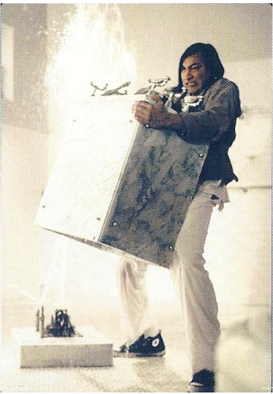 A man with dark hair and light-colored pants holding a painting with a small sculpture on a table in front of him, in a bright room.