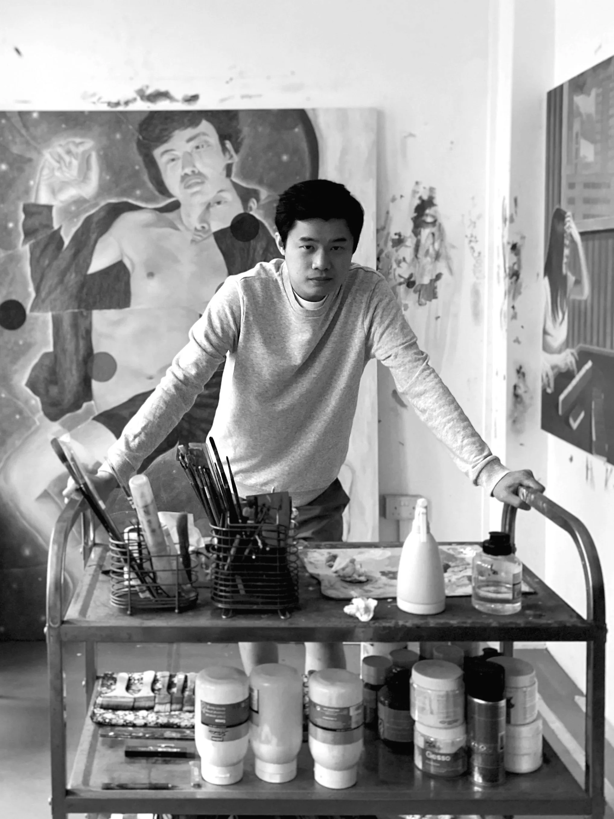 A young man with dark hair wearing a light-colored long-sleeve shirt standing behind a rolling art cart, looking at the camera. There are art supplies such as brushes, bottles, and jars on the cart. A large painting of a man with an open shirt and a raised fist looms in the background, with another partial painting visible on the wall to the right.