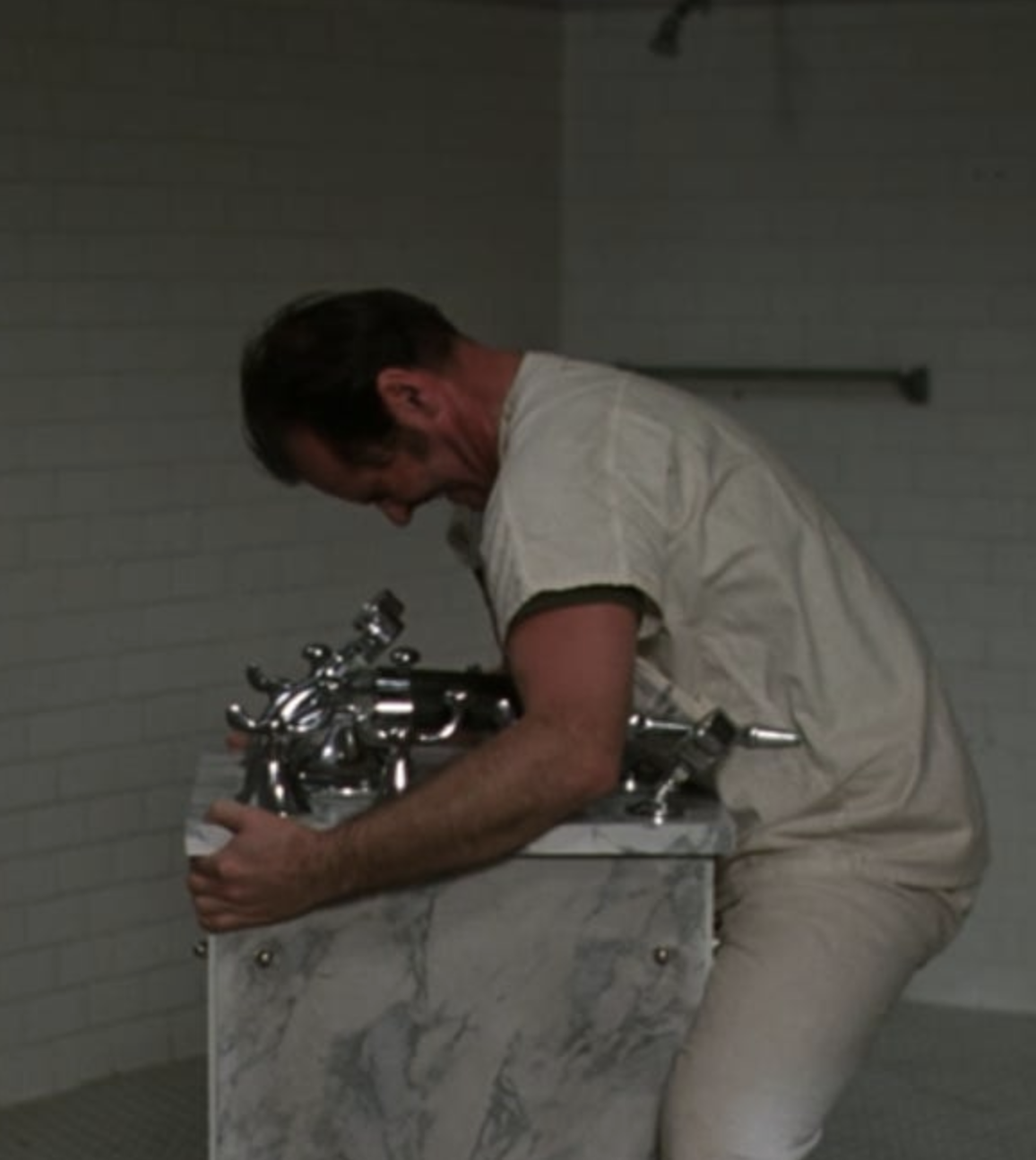A man leaning over a marble sink, holding metal plumbing fixtures, in a room with white brick walls.