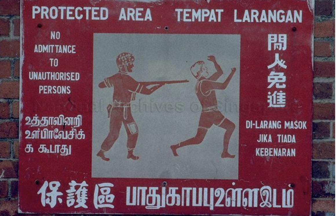 A warning sign on a brick wall with text in multiple languages, including English, Chinese, Tamil, and Malay, indicating a protected area where unauthorized persons are not allowed. The sign features an illustration of a person aiming a gun at another person, who is raising their hands in surrender.