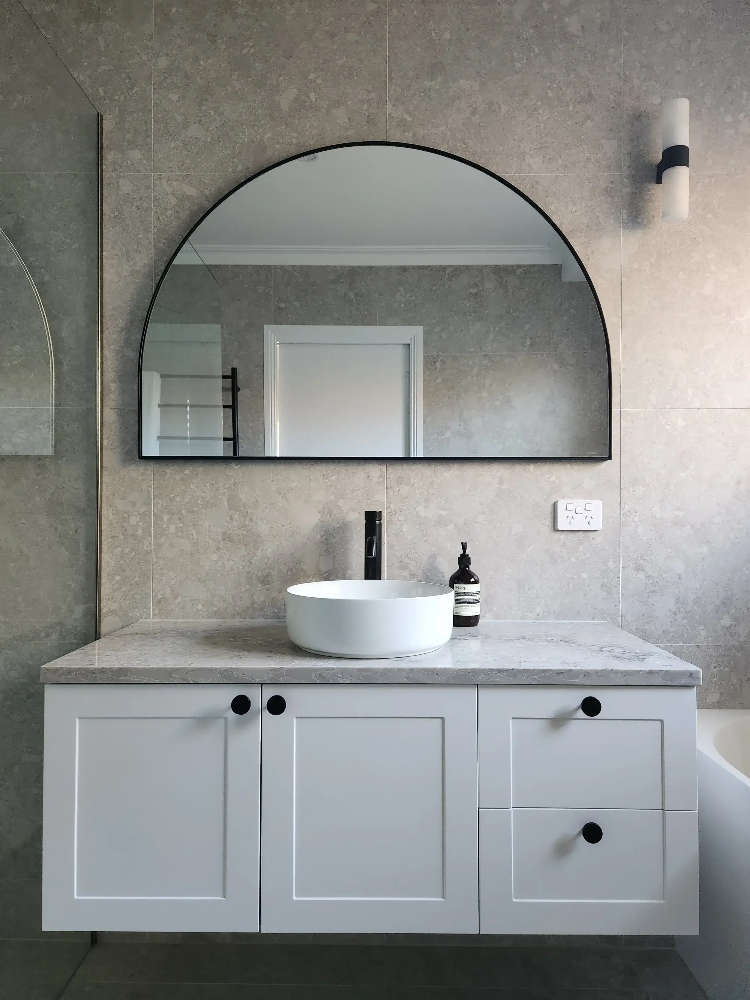 Modern bathroom vanity area with a white cabinet, grey marble countertop, round vessel sink, black faucet, and a large arched mirror. There's a soap dispenser and electrical outlets on the wall, with a textured grey tile wall and a white door.