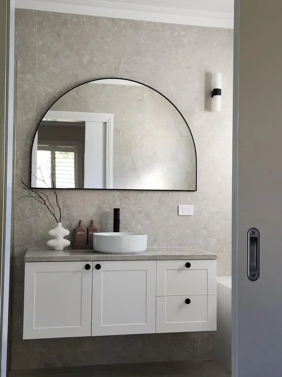 Modern bathroom with a wall-mounted mirror, white vanity with black knobs, vessel sink, black faucet, decorative vase with branches, soap dispensers, and a wall-mounted light fixture.