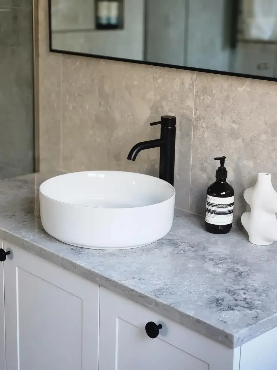 Bathroom vanity with a white vessel sink, black faucet, a black soap dispenser, and a white decorative vase, with a large mirror on a beige tiled wall.