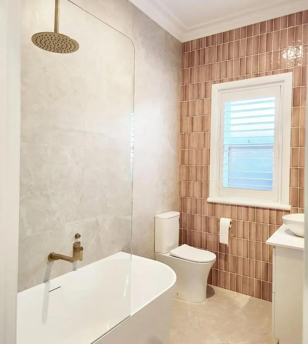 Bathroom with a bathtub, a toilet, a window with shutters, and pink tile walls.