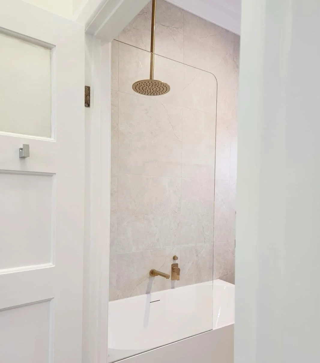 View of a shower with a brass rain shower head, a transparent glass shower barrier, and beige tiled walls, seen through a partially open white door.
