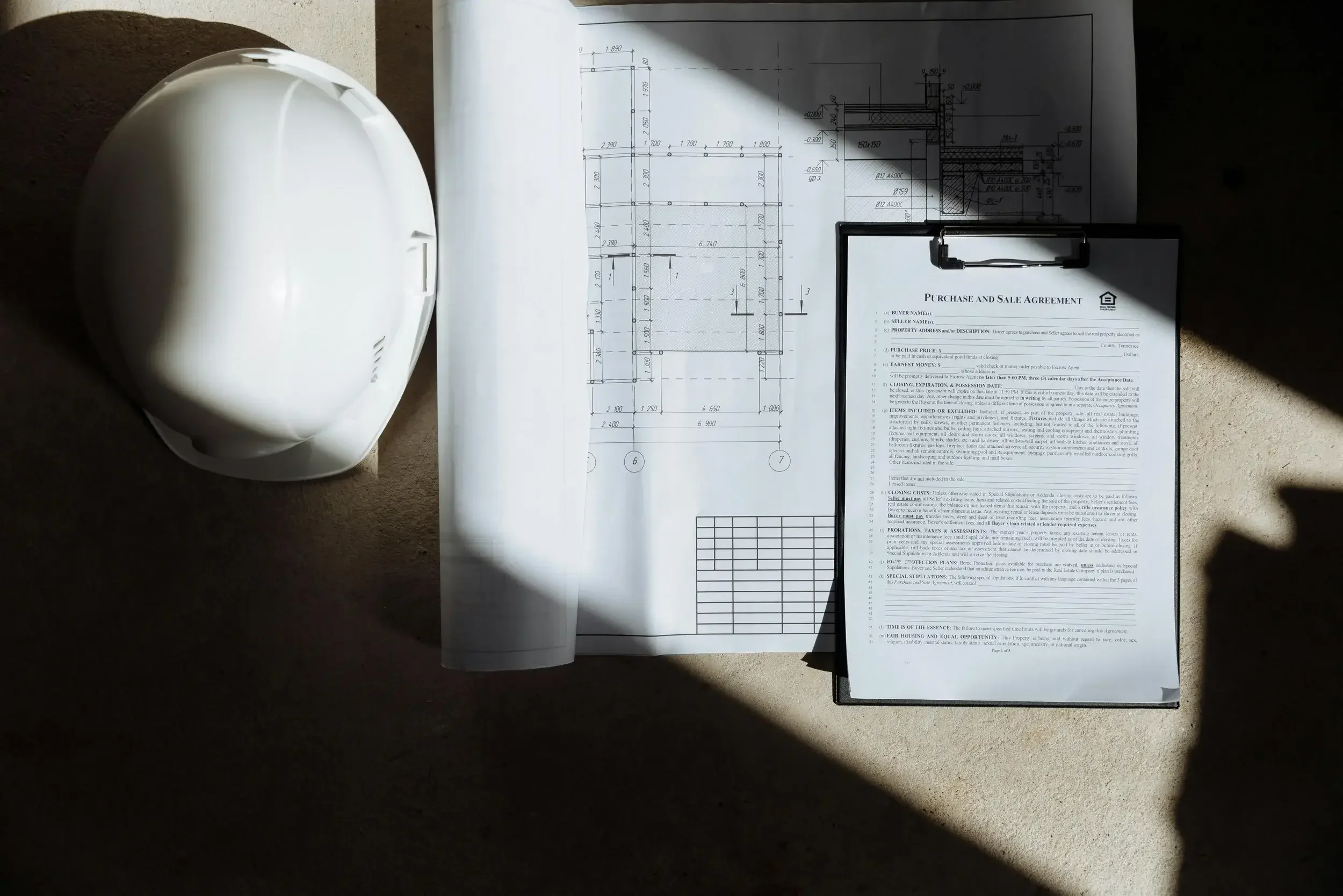 A white construction helmet, a rolled architectural blueprint, and a clipboard with a purchase and sale agreement resting on a beige surface, with sunlight casting shadows.