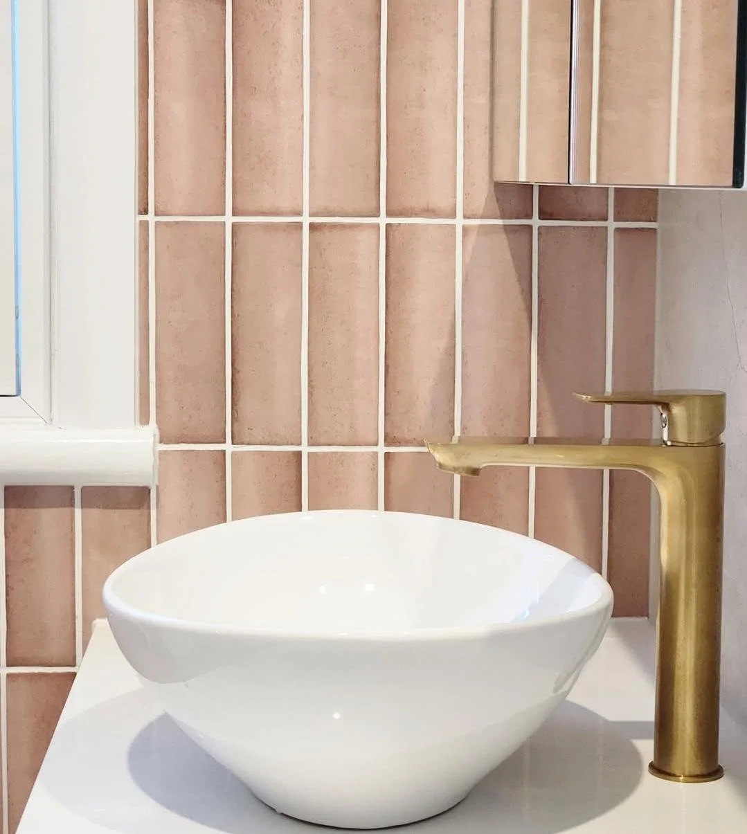 A white bowl-shaped vessel with a matte gold faucet on a bathroom countertop, against pink tiled walls.