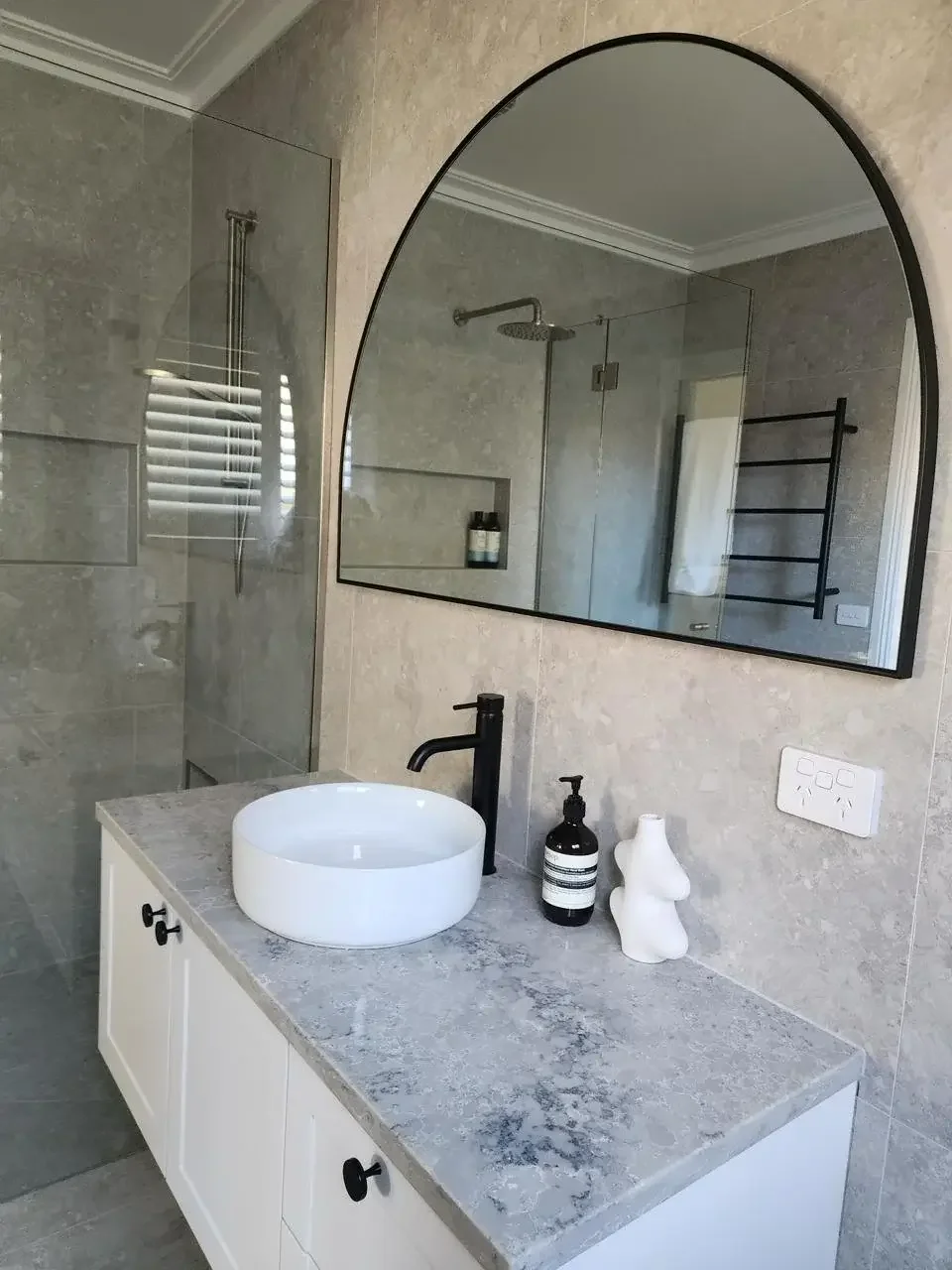 Modern bathroom with gray stone countertop, white vessel sink, black faucet, decorative white sculpture, dark soap dispenser, large arched mirror, shower area with glass door, towel on black ladder rack, and window with shutters.