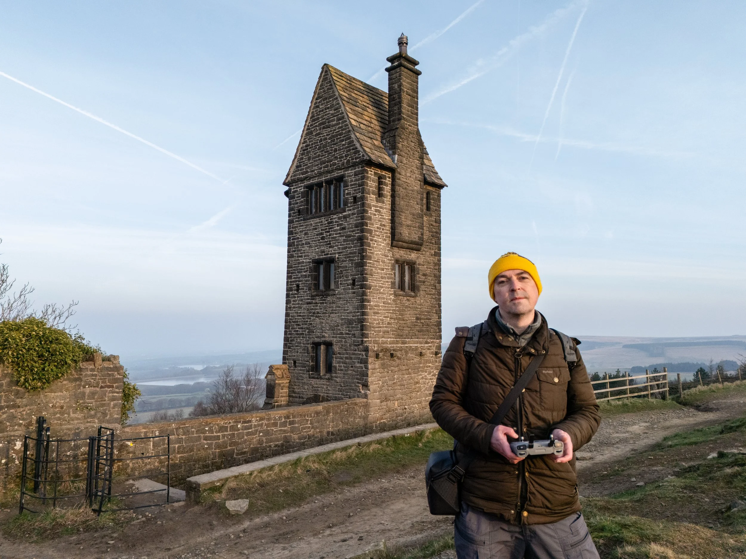 Man with a yellow beanie and brown jacket holding a remote control, standing outdoors in front of a stone tower with a pointed roof, overlooking a landscape with hills and water.