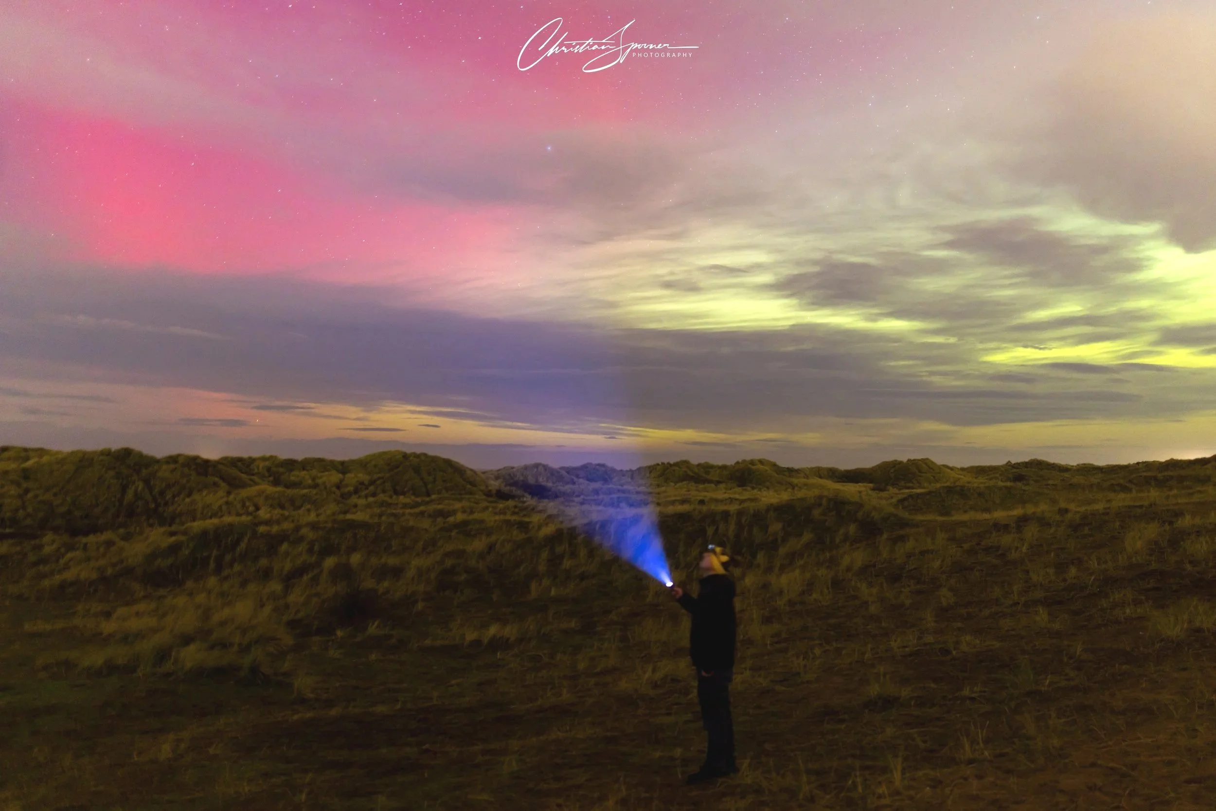 Person holding a flashlight in a grassy landscape during night, illuminating the sky with northern lights displaying pink, green, and purple hues with stars.
