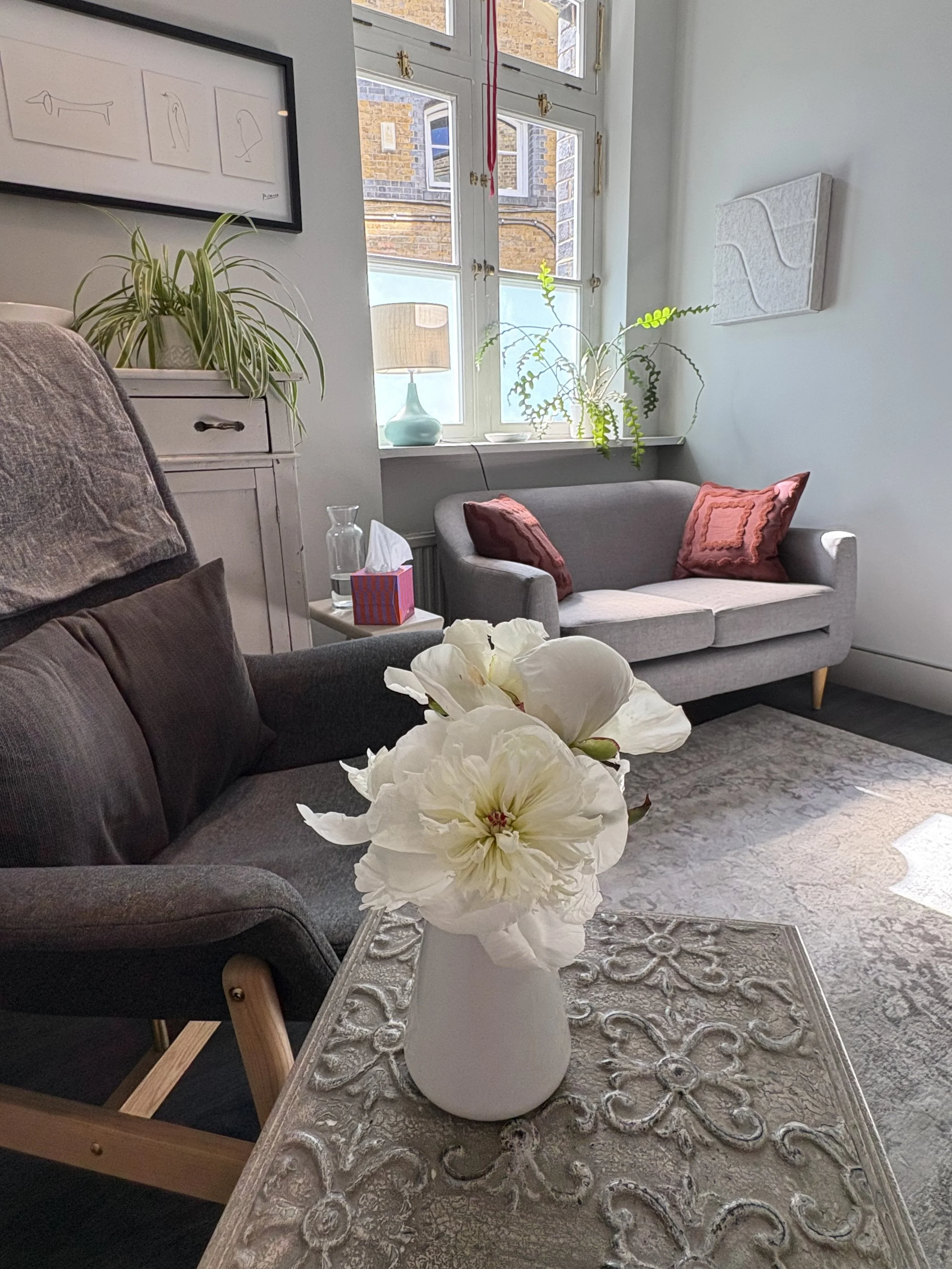 A calm, cosy and bright therapy room with a comfy sofa, a side table with a jug of water and a vase with fresh flowers. There is a window letting in sunlight, there are plants, artwork, and an armchair.