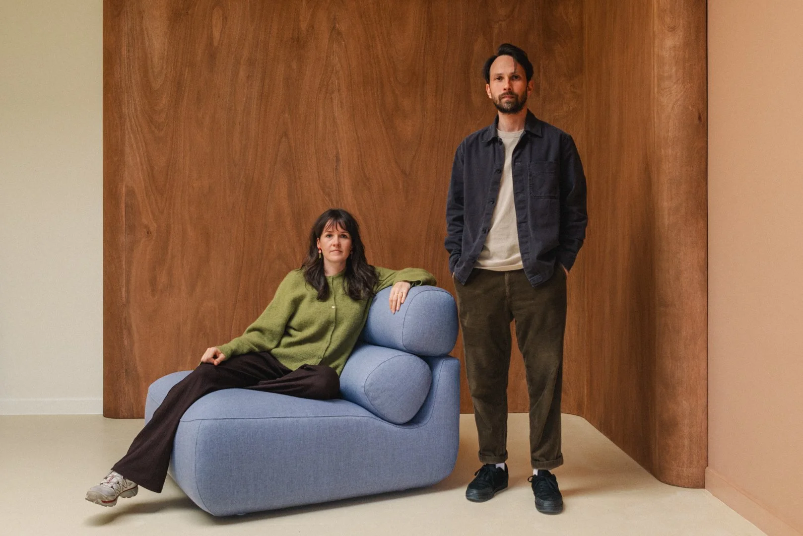 Een man en een vrouw in een moderne interieurruimte. De vrouw zit op een lichtblauwe bank, gekleed in een groene trui, en de man staat ernaast met zijn handen in zijn zakken, gekleed in een donkere jas en bruine broek. Achter hen een houten muur.