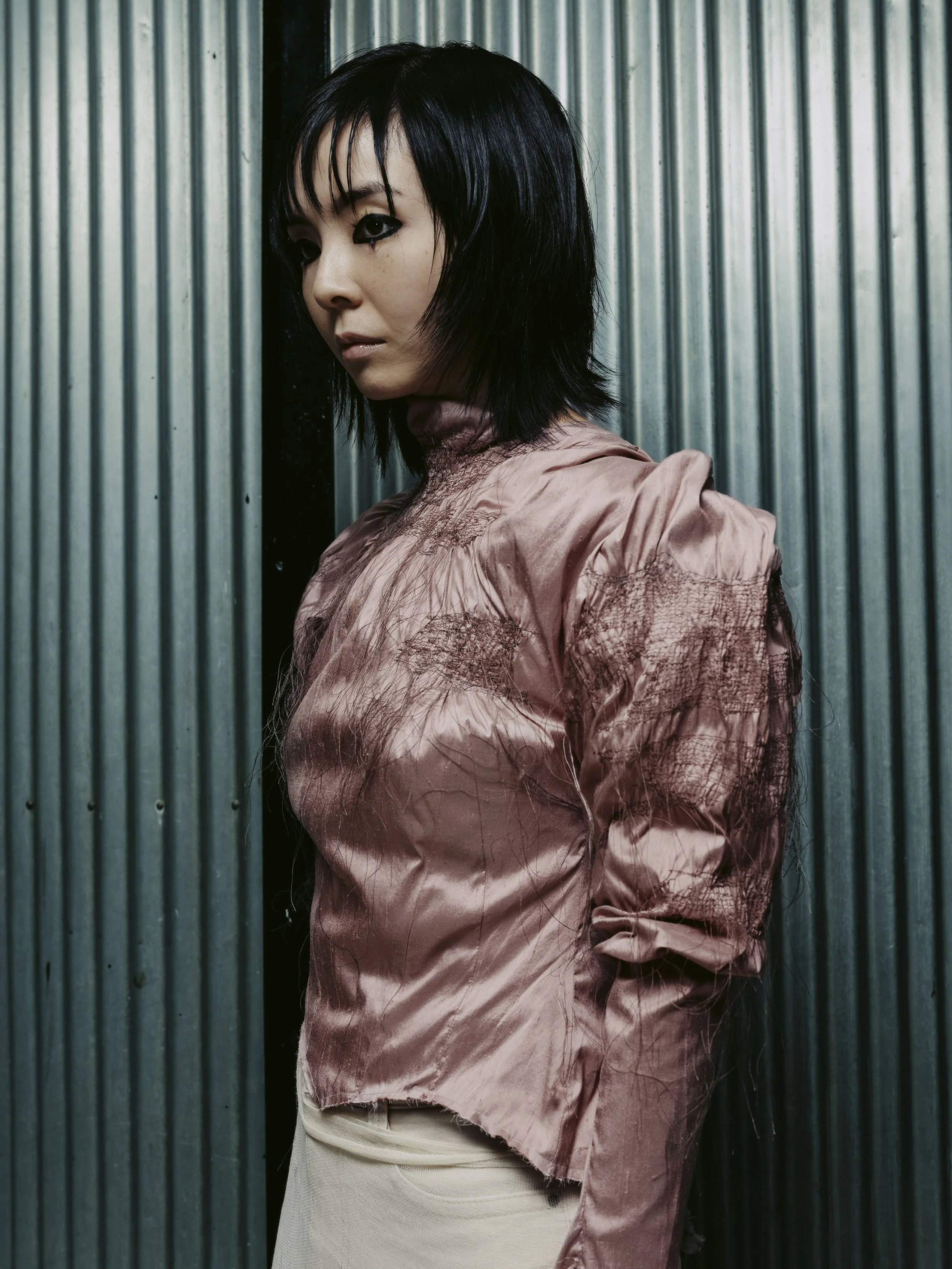 Porträt einer asiatischen Frau mit schwarzem, gestyltem Haar, die an einer Wand mit vertikalen Metallblechpaneelen lehnt.