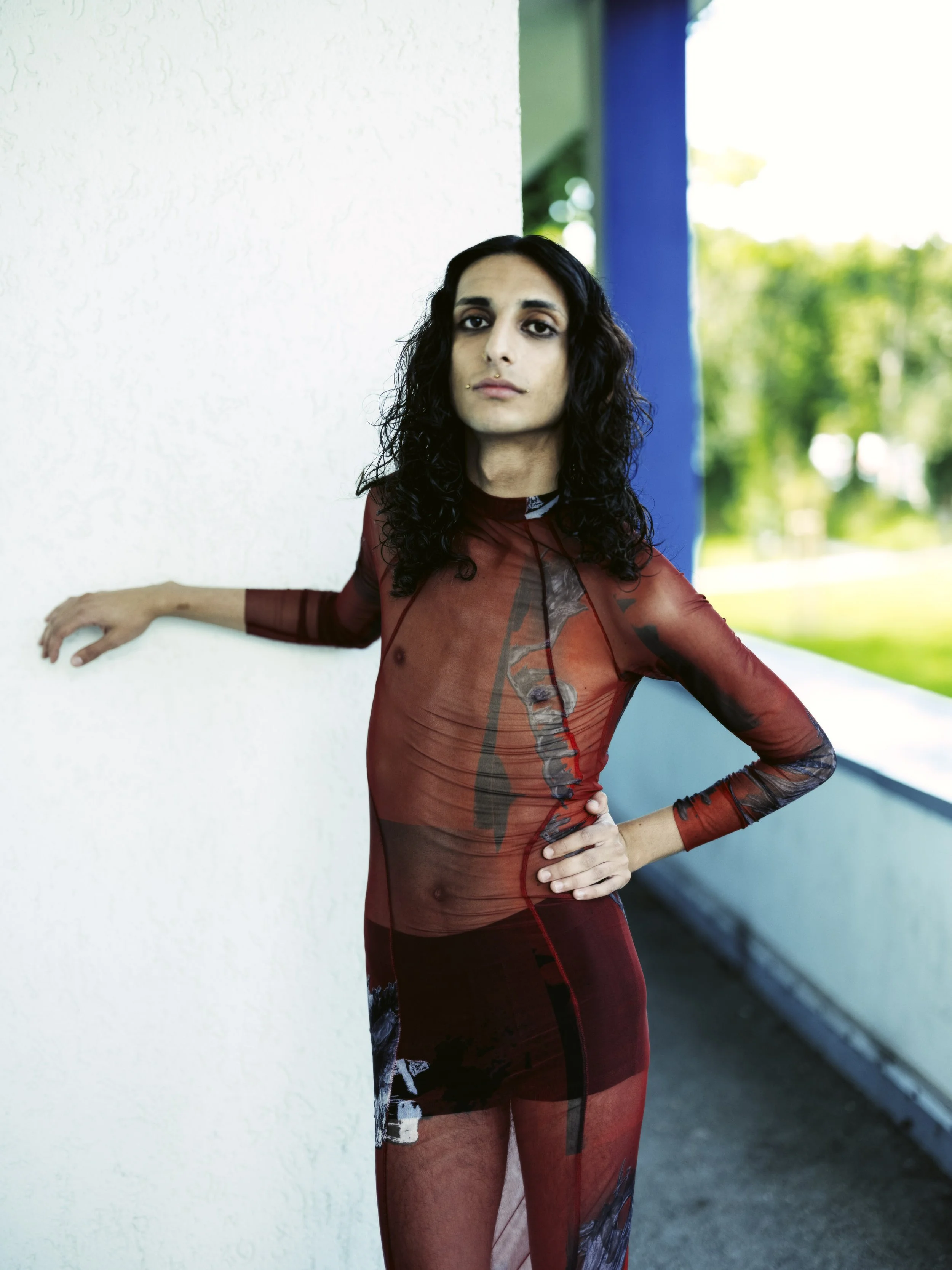 Porträt einer Person mit langer, dunkler, lockiger Haarfrisur, die ein transparentes rotes Kleid trägt, das Muster aufweist, und an einer weißen Wand lehnt, im Hintergrund grüner Garten bei Tag.
