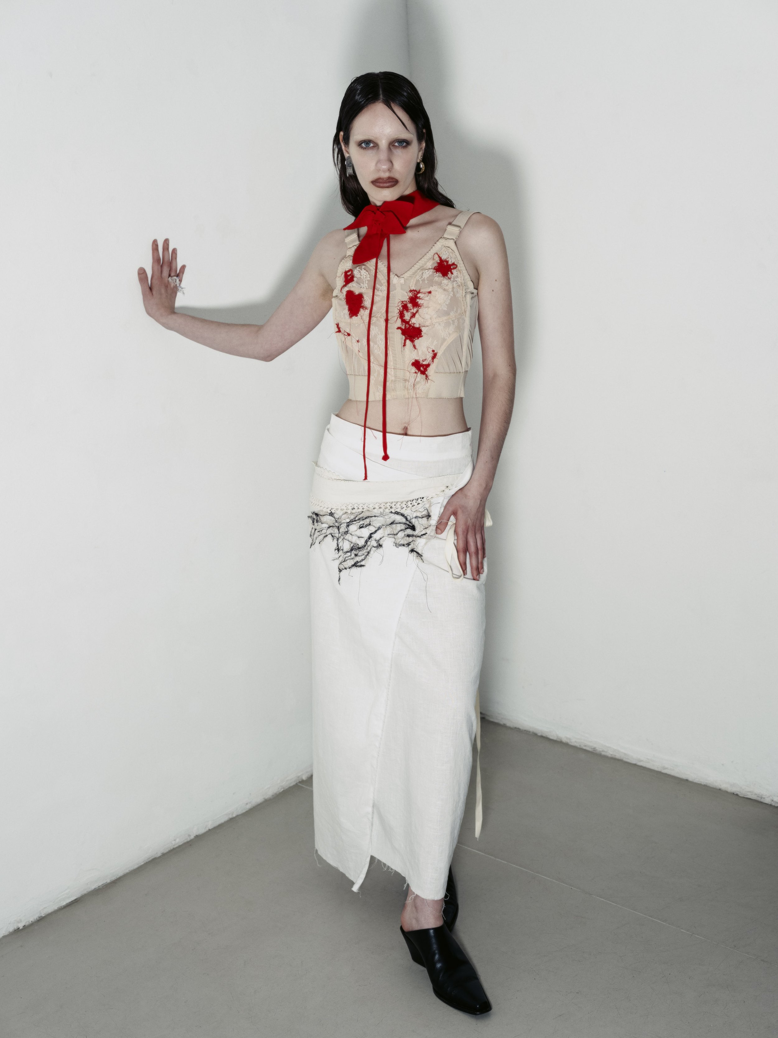Eine junge Frau steht in einem weißen Raum, trägt ein auffälliges, kreatives Outfit mit roten Stickereien auf einem oberen, beige farbenen Top, einer langen weißen Rock mit schwarzen Verzierungen, schwarzen Schuhen und einem roten Band um den Hals.