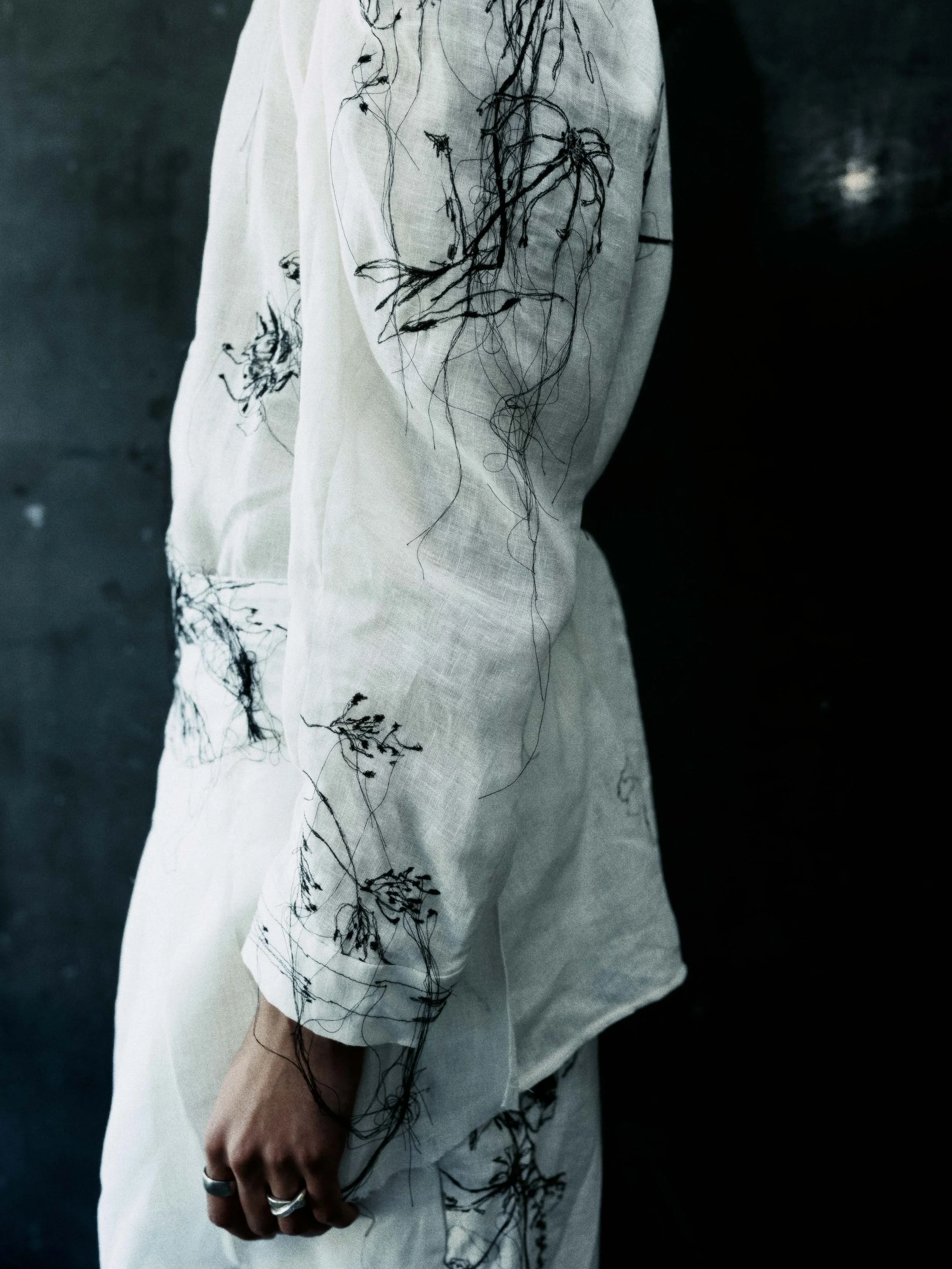 Nahaufnahme einer Person in einem weißen Oberteil mit schwarzen Stickereien, die wie feine filigrane Äste oder Wurzeln aussehen. Die Person trägt Ringe an der Hand und steht vor dunklem Hintergrund.