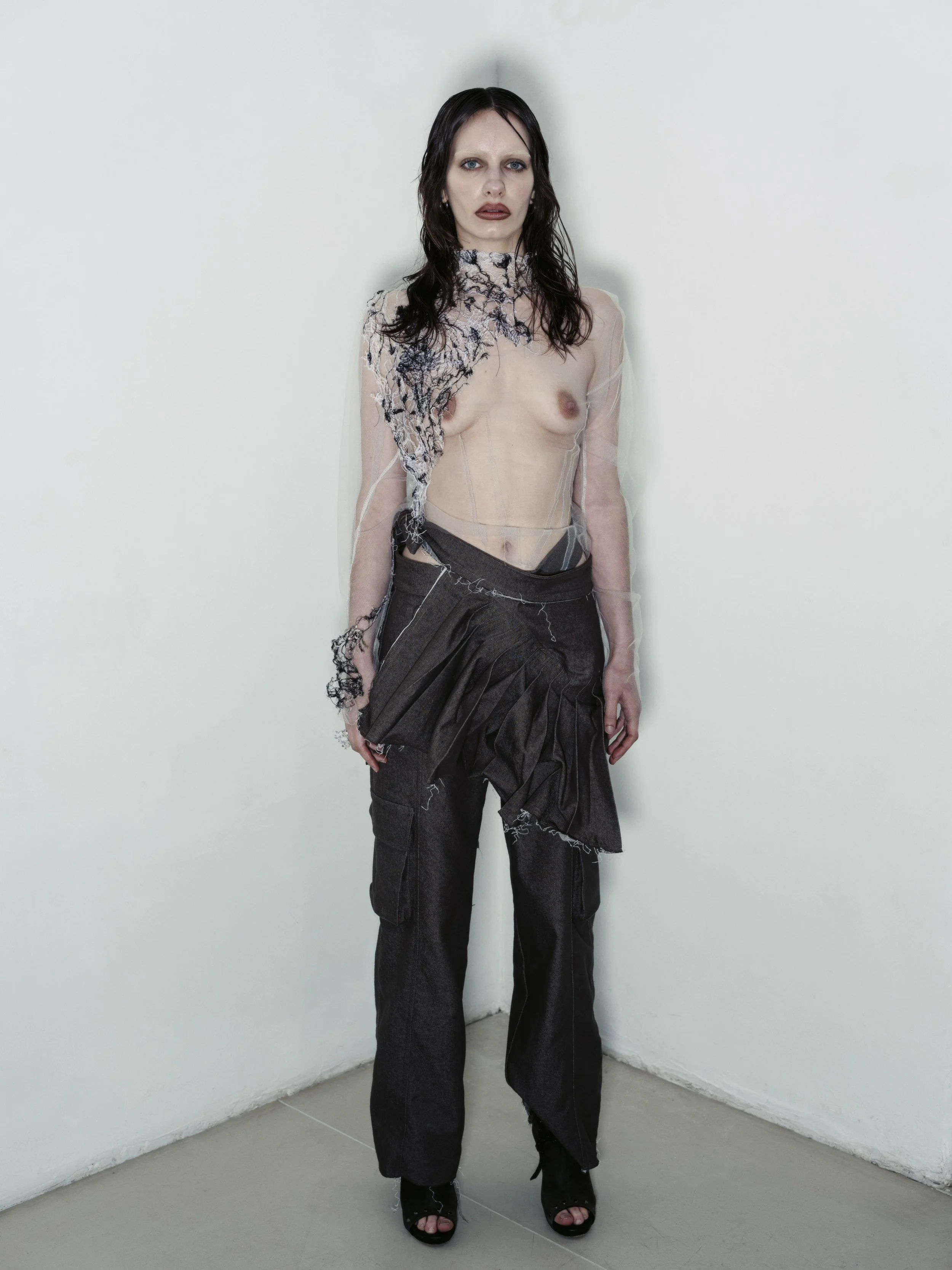 Junge Frau mit dunklen, nassen Haaren steht in einem weißen Raum, trägt keine Oberbekleidung, nur eine durchsichtige, verzierte Oberfläche und dunkle Hosen.