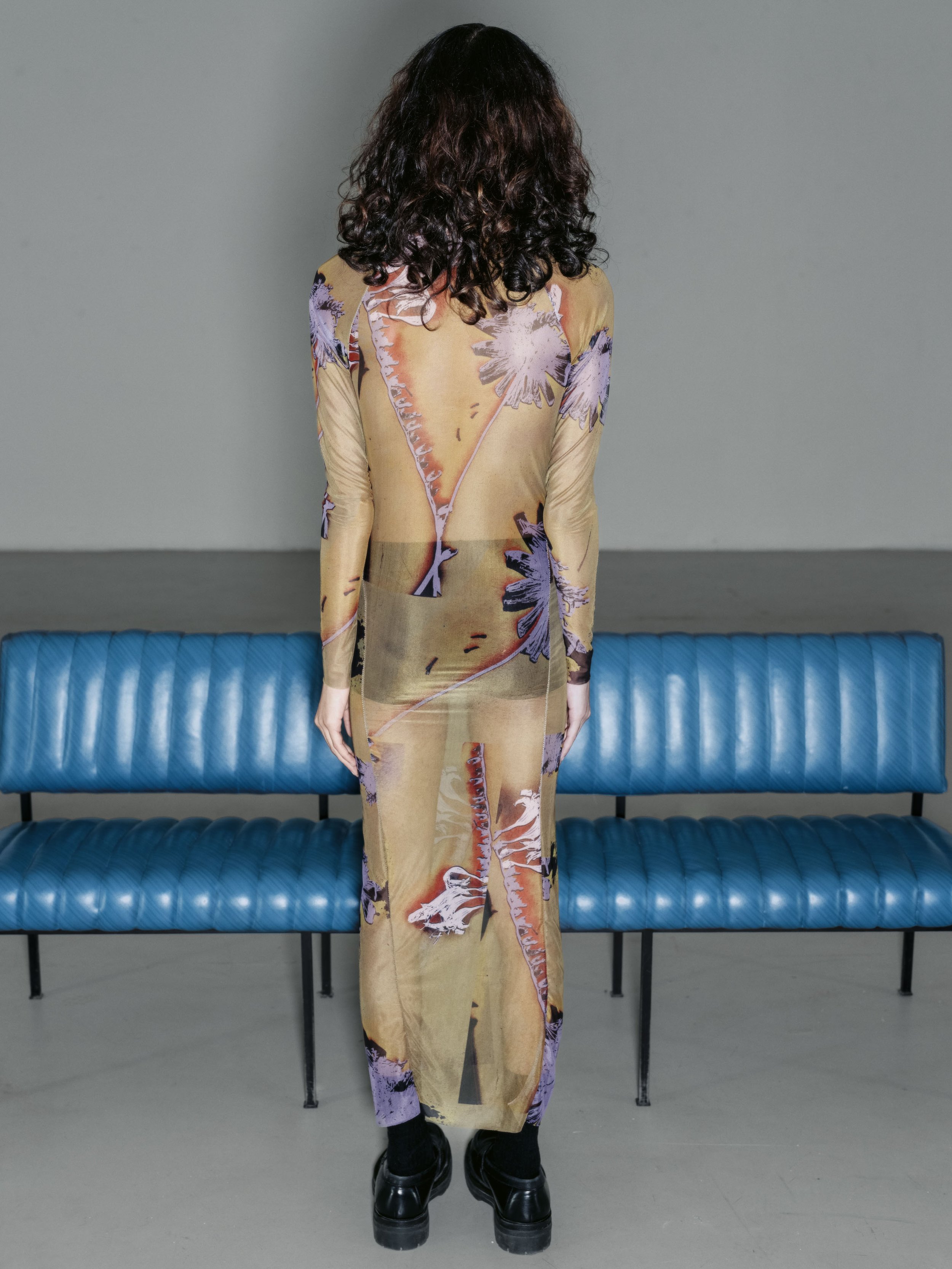 Frau steht vor einer blauen Bank, trägt ein langes, transparentes Kleid mit Blumenmuster, Sicht auf Rücken und Beine