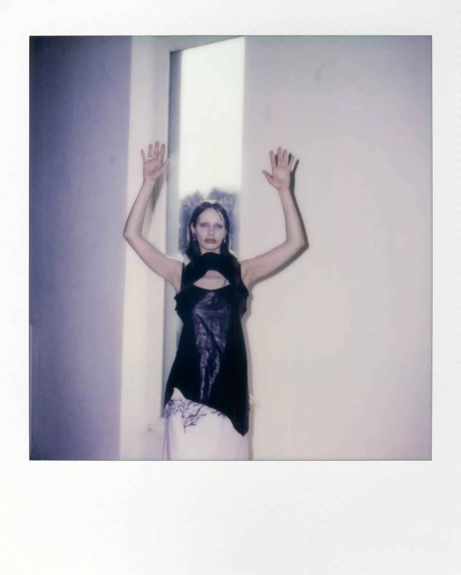 Eine junge Frau in einem schwarzen Kleid steht mit erhobenen Armen vor einem weißen Wand und einem Fenster, aus dem Tageslicht scheint.