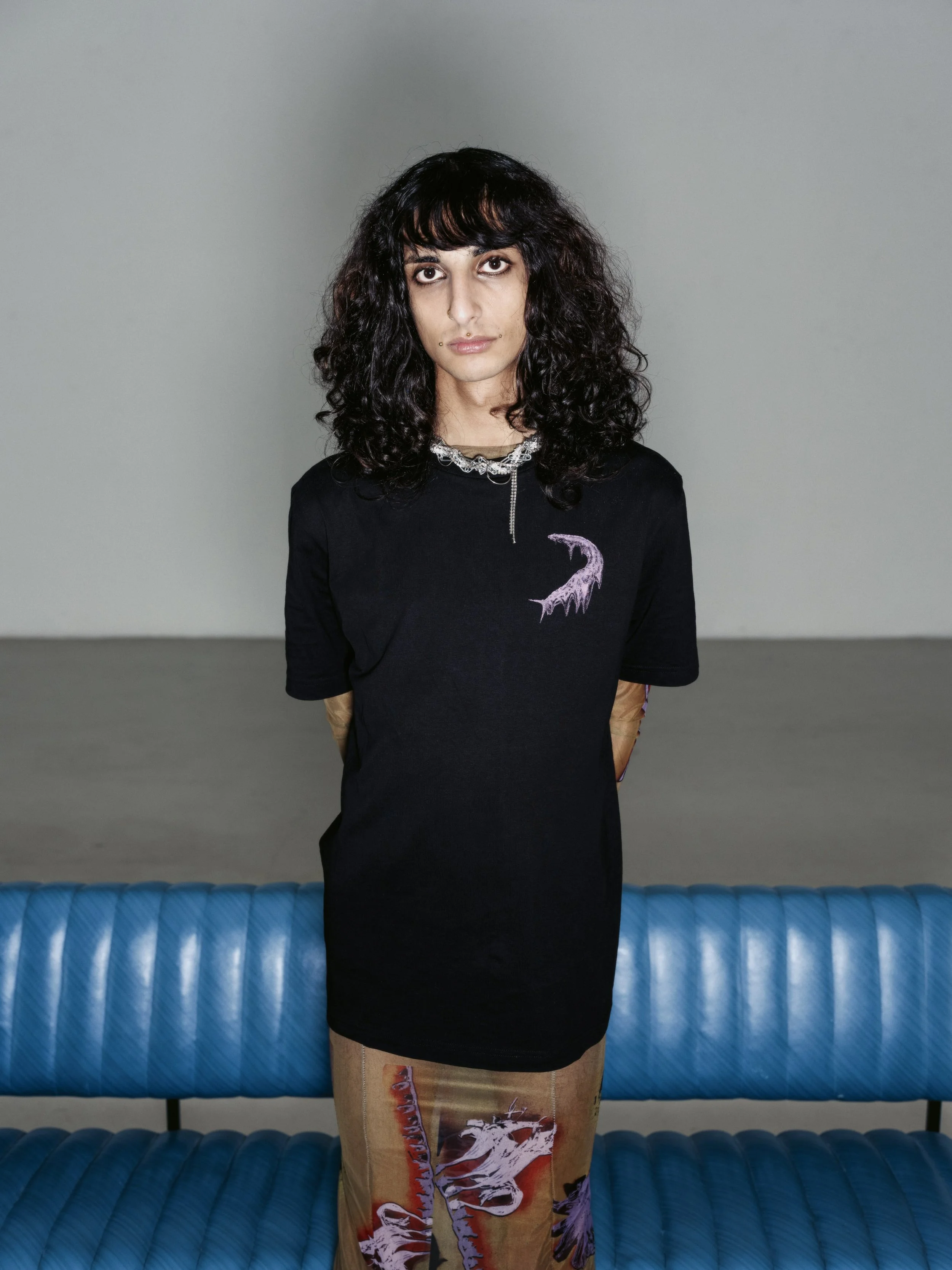 Eine Frau mit dunklen, lockigen Haaren steht vor einem grauen Hintergrund. Sie trägt ein schwarzes T-Shirt mit einem lilafarbenen Seeungeheuer-Design und bunte Hosen mit Dinosaurier-Druck.