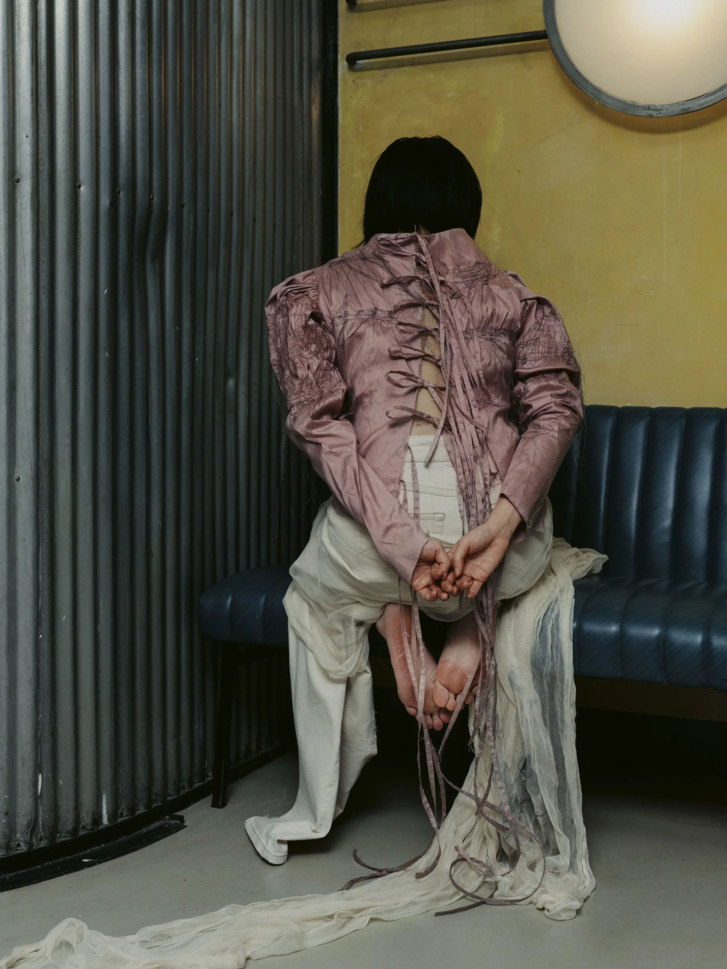 Eine Person sitzt auf einer Bank, trägt eine ausgewaschene, ruchige Jacke und eine lang fließende Hose. Die Person ist von hinten zu sehen, die Hände sind hinter dem Rücken verschränkt, und die Beine sind über den Knien übereinander geschlagen.