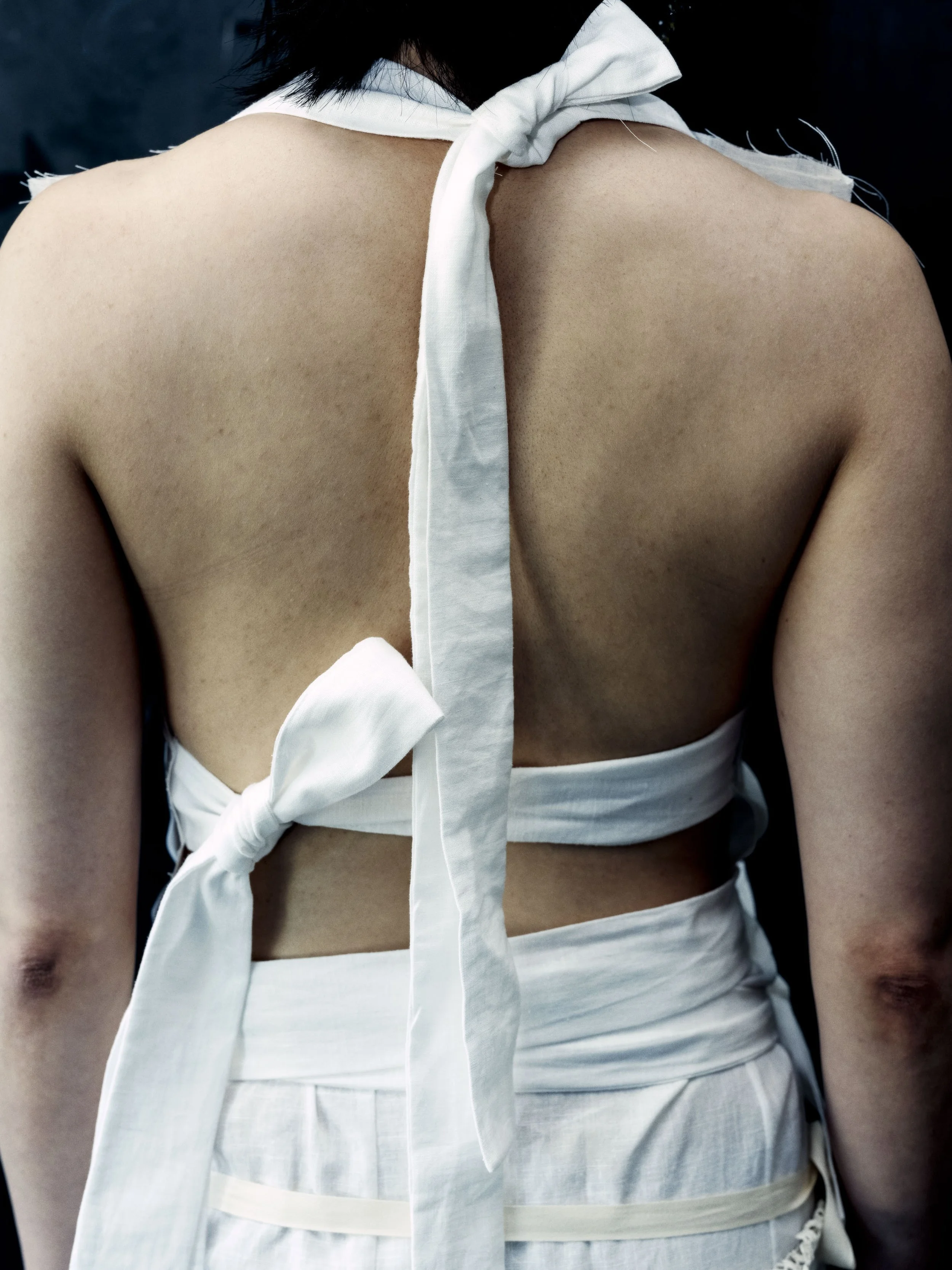 Nahaufnahme des Rückens einer Person, die weißes Stoffkleid trägt, das vorne zugebunden ist.