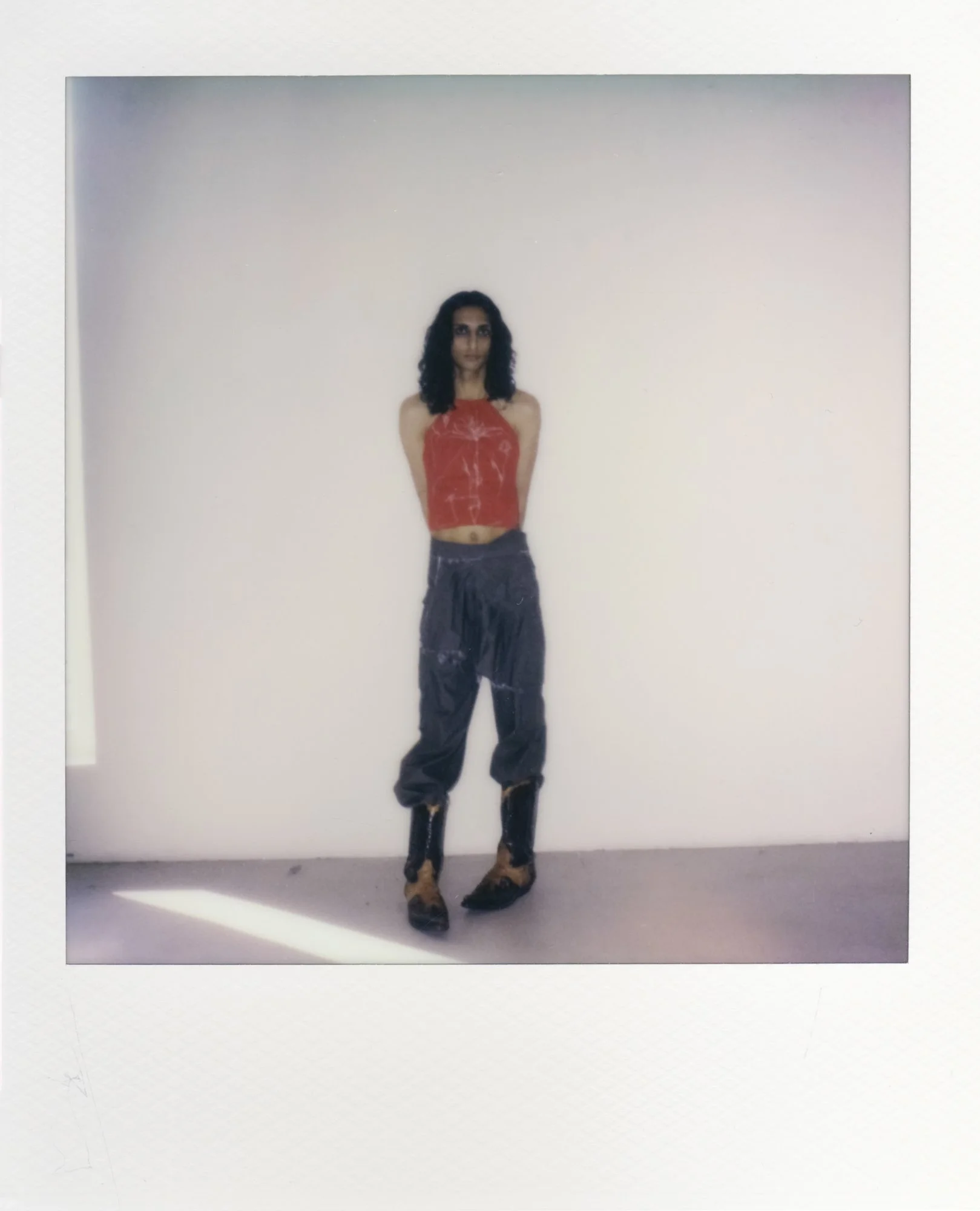 Frau in rotem Top, schwarzen Hosen und Stiefeln steht vor weißer Wand in einem weißen Raum.