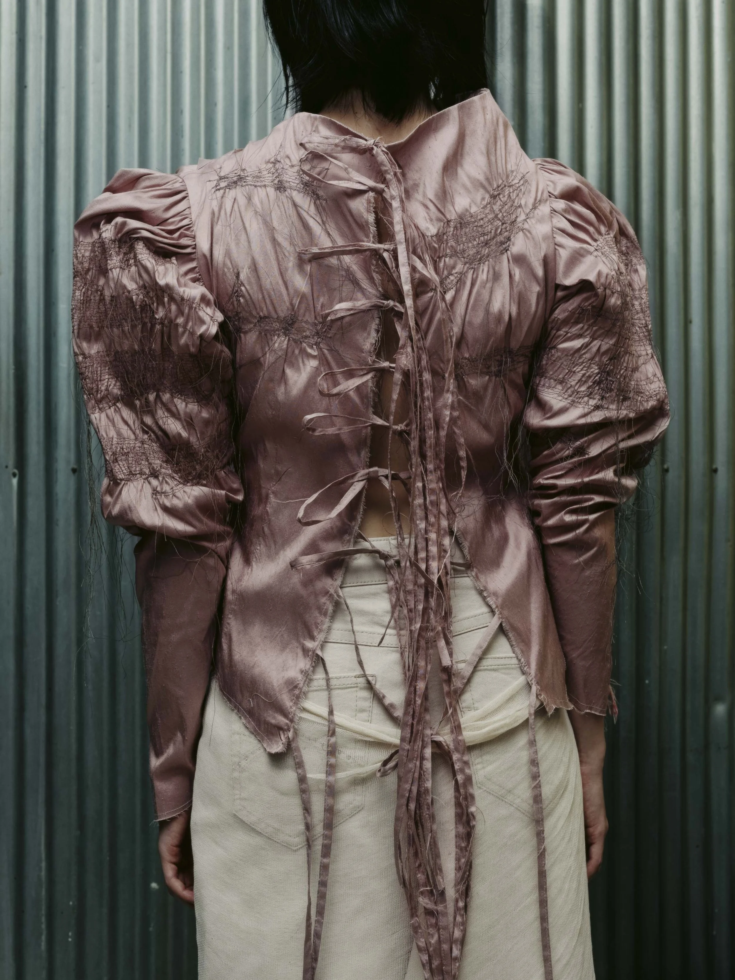 Eine Person trägt eine rosa, verzierte Bluse vor einer metallischen Wand.
