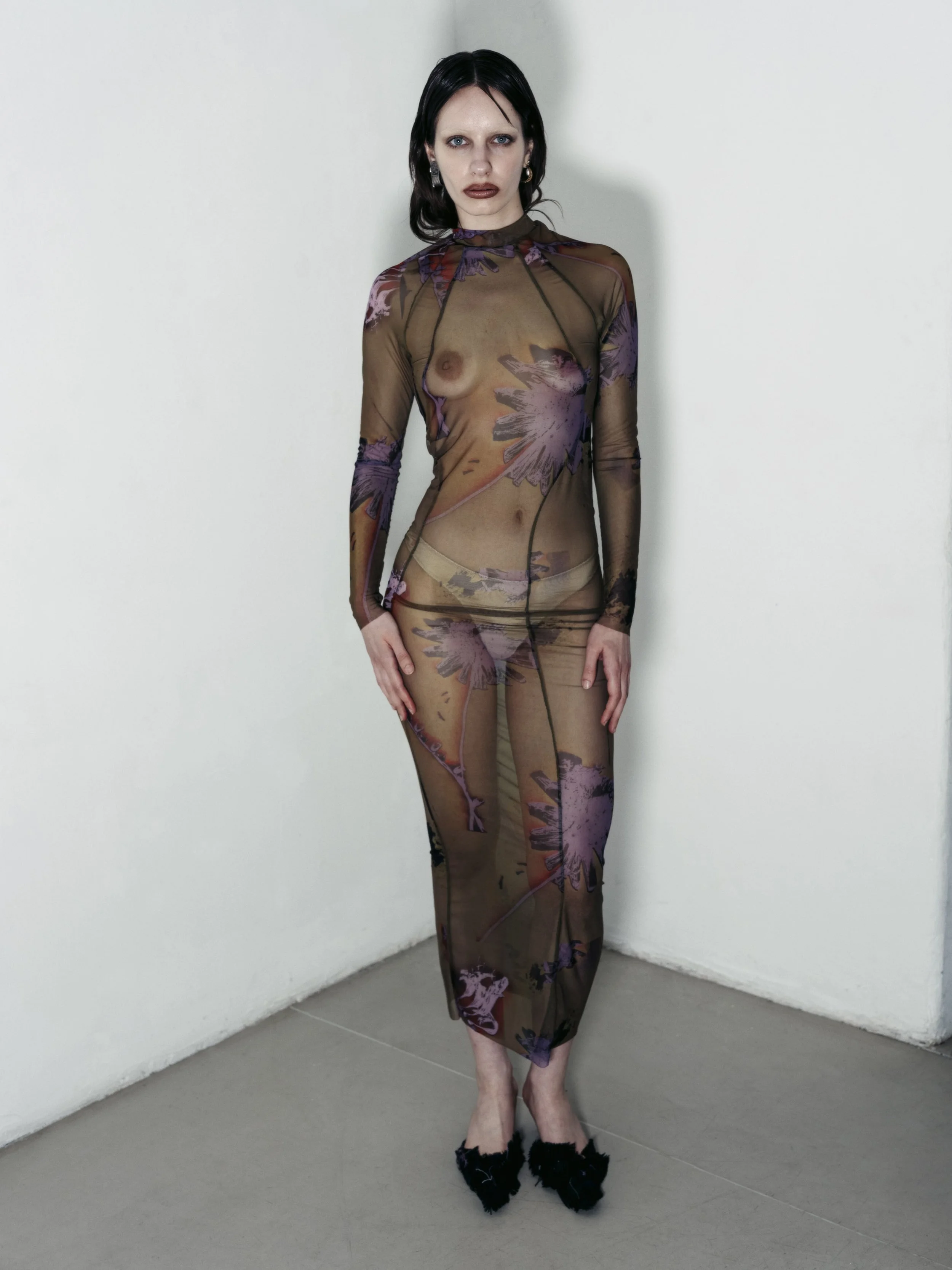 Eine Frau in einem transparenten, langärmligen Kleid mit Blumenmuster steht in einer Ecke eines Raumes.