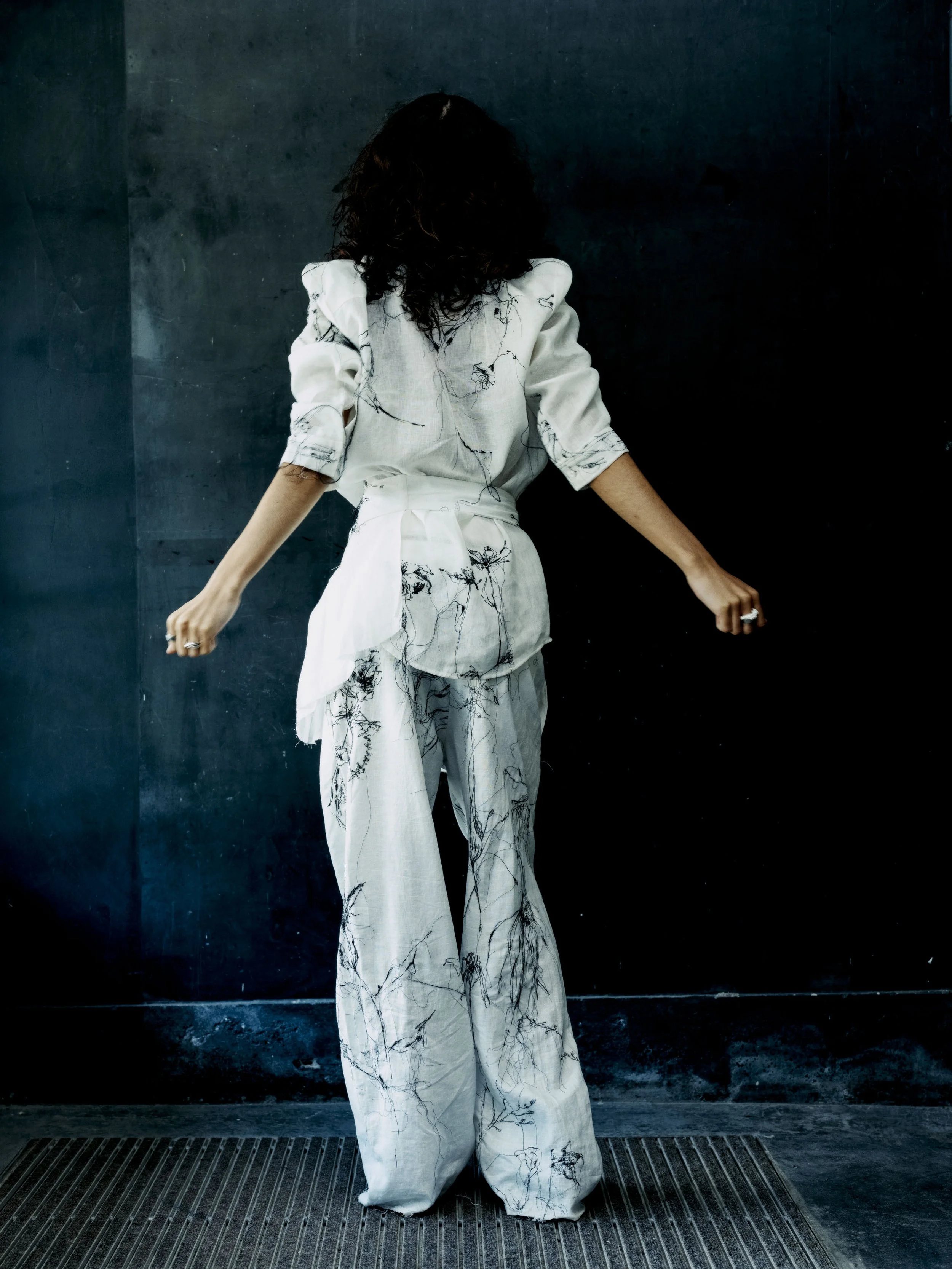 Frau in weißem, gemustertem Anzug steht vor dunkler Wand, den Rücken zum Betrachter, Arme seitlich ausgestreckt.