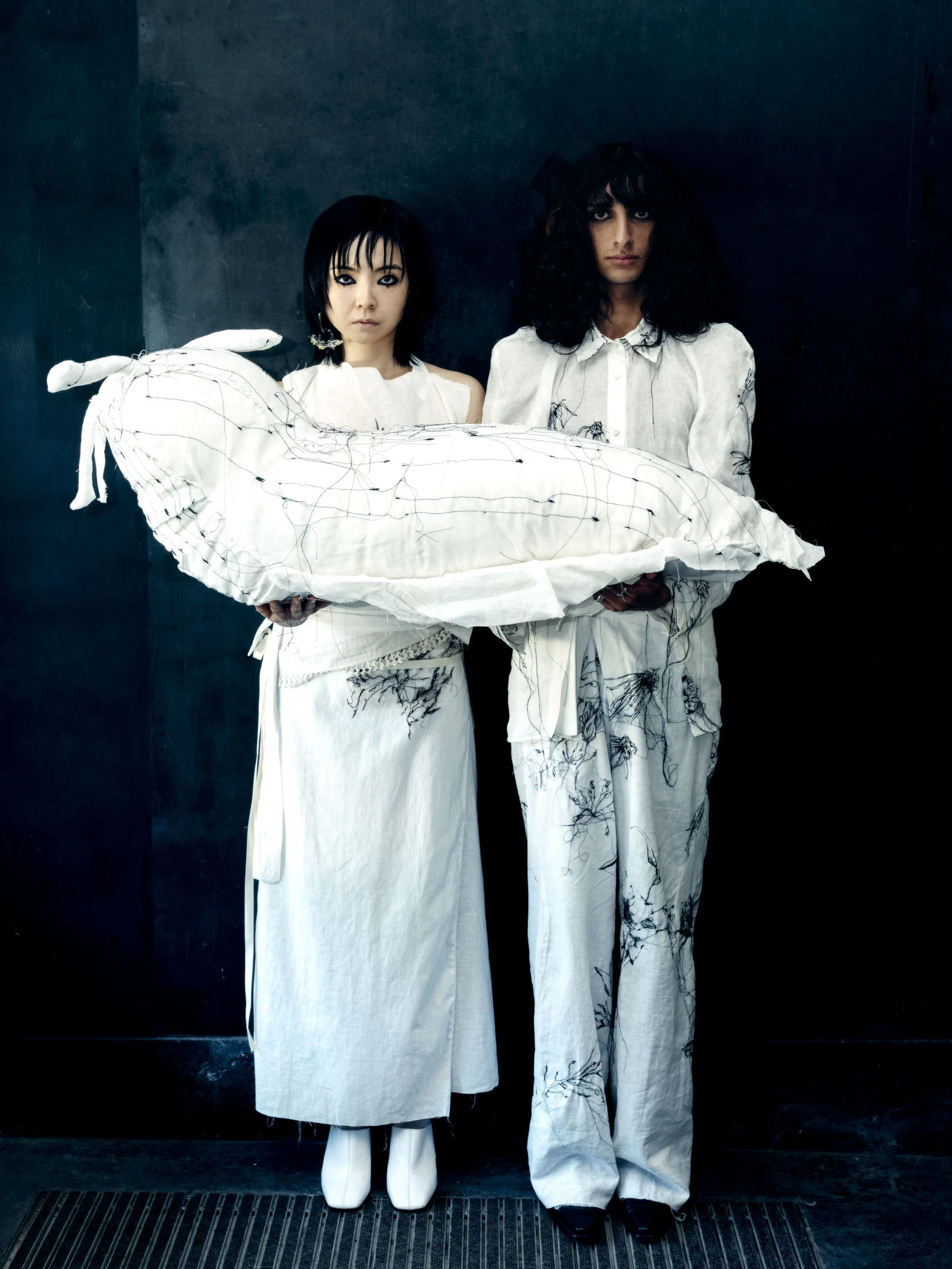 Zwei Menschen in weißen Kleidern stehen vor einem schwarzen Hintergrund, die eine trägt weiße Schuhe, die andere schwarze, und sie halten gemeinsam eine große, weiße, schädelartige Skulptur.