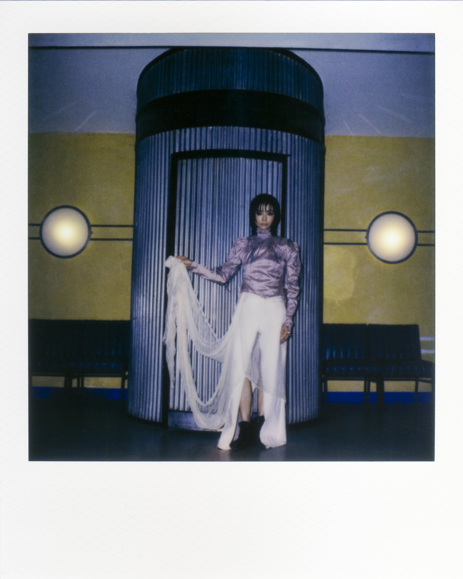 Eine Frau in schicker Kleidung steht vor einer Tür in einem modernen Raum, die sie mit einer weißen Stoffbahn in der Hand hält.