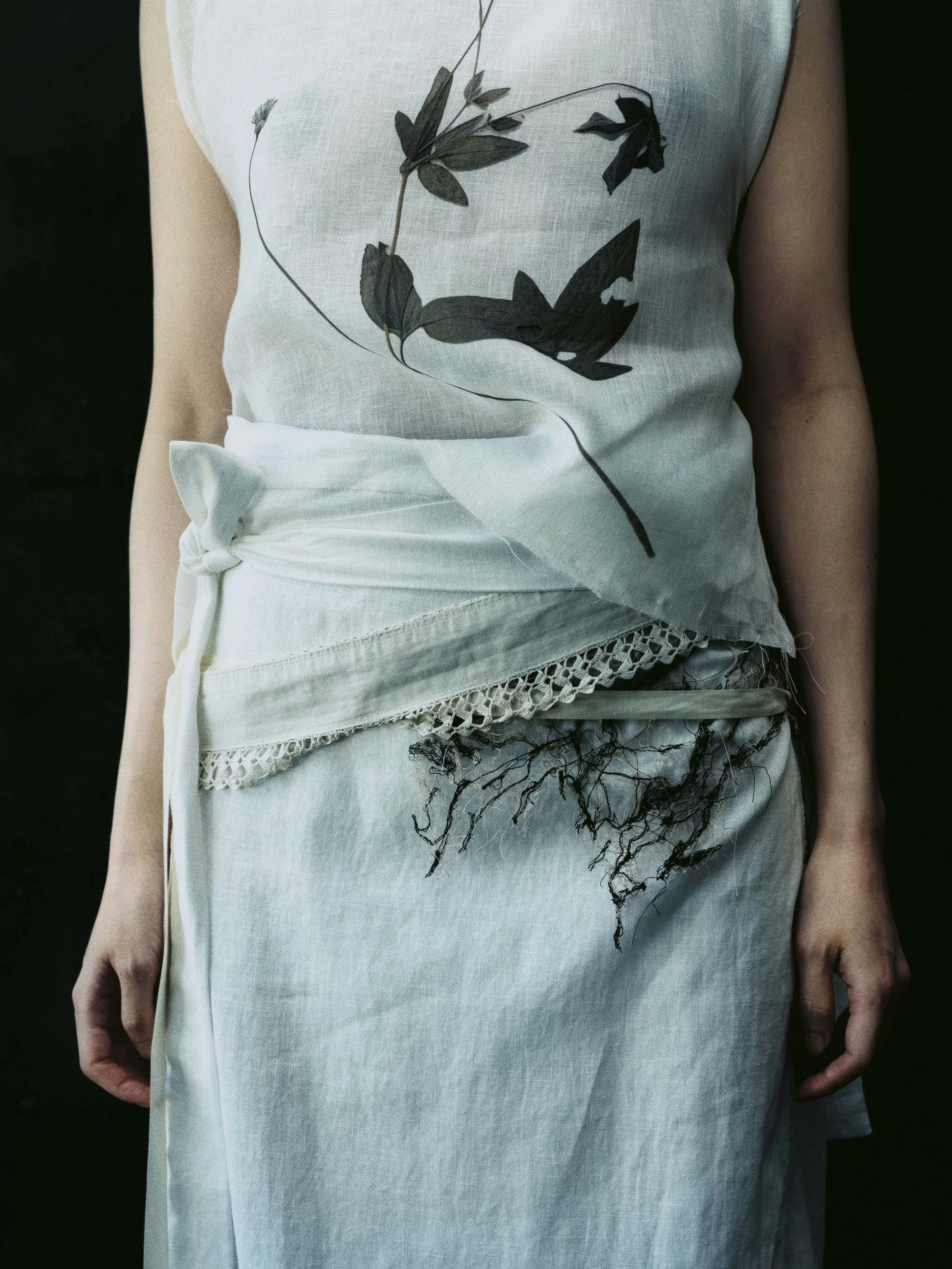 Person in weißem Kleid mit floralem Druck, der an einer Seite gebunden ist und Spitze am Saum hat, steht vor dunklem Hintergrund.