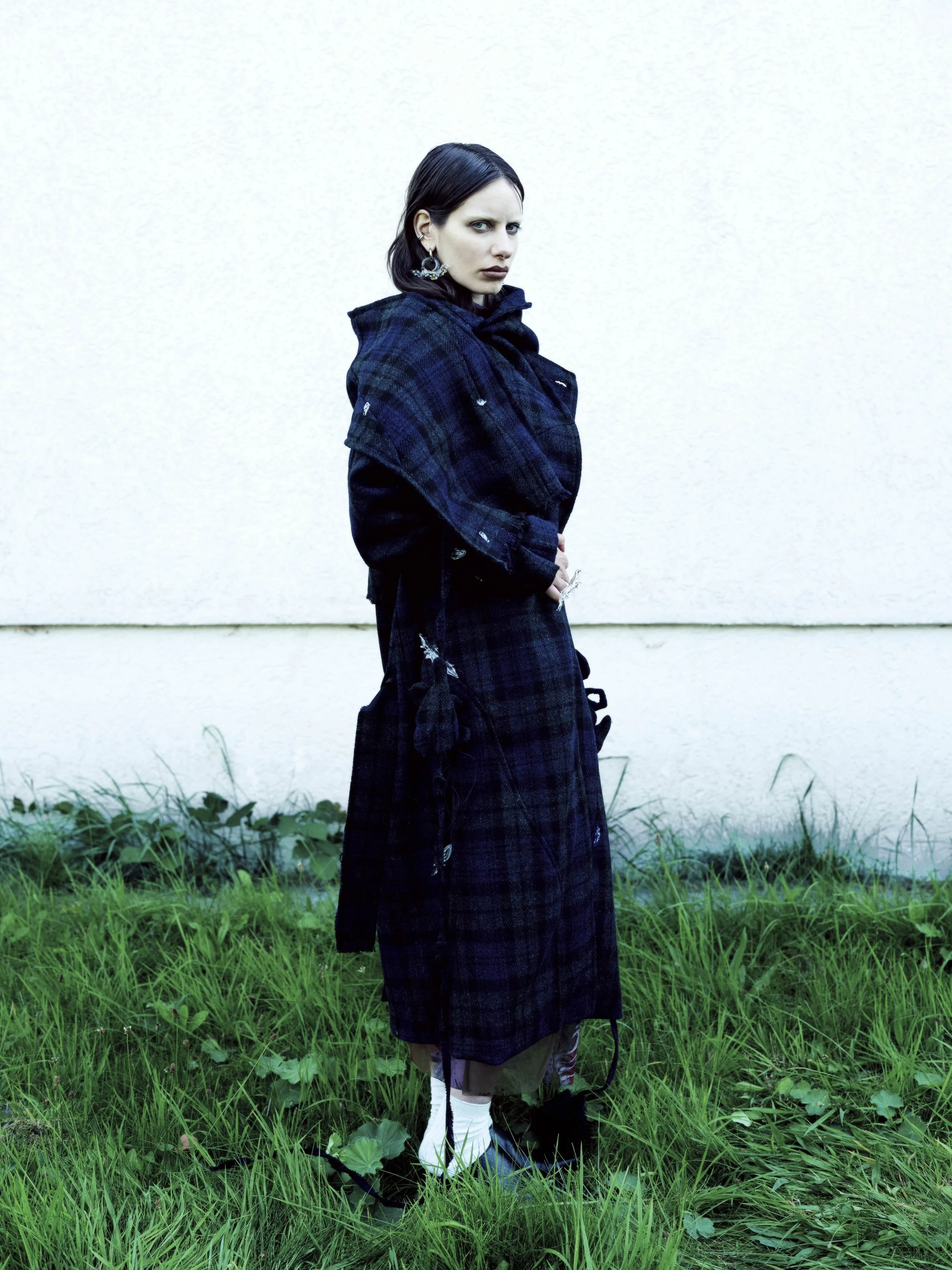 Eine Frau mit schwarzen Haaren in dunkler, kariert Kleidung steht auf grünem Gras vor einer weißen Wand, schaut ernst in die Kamera.