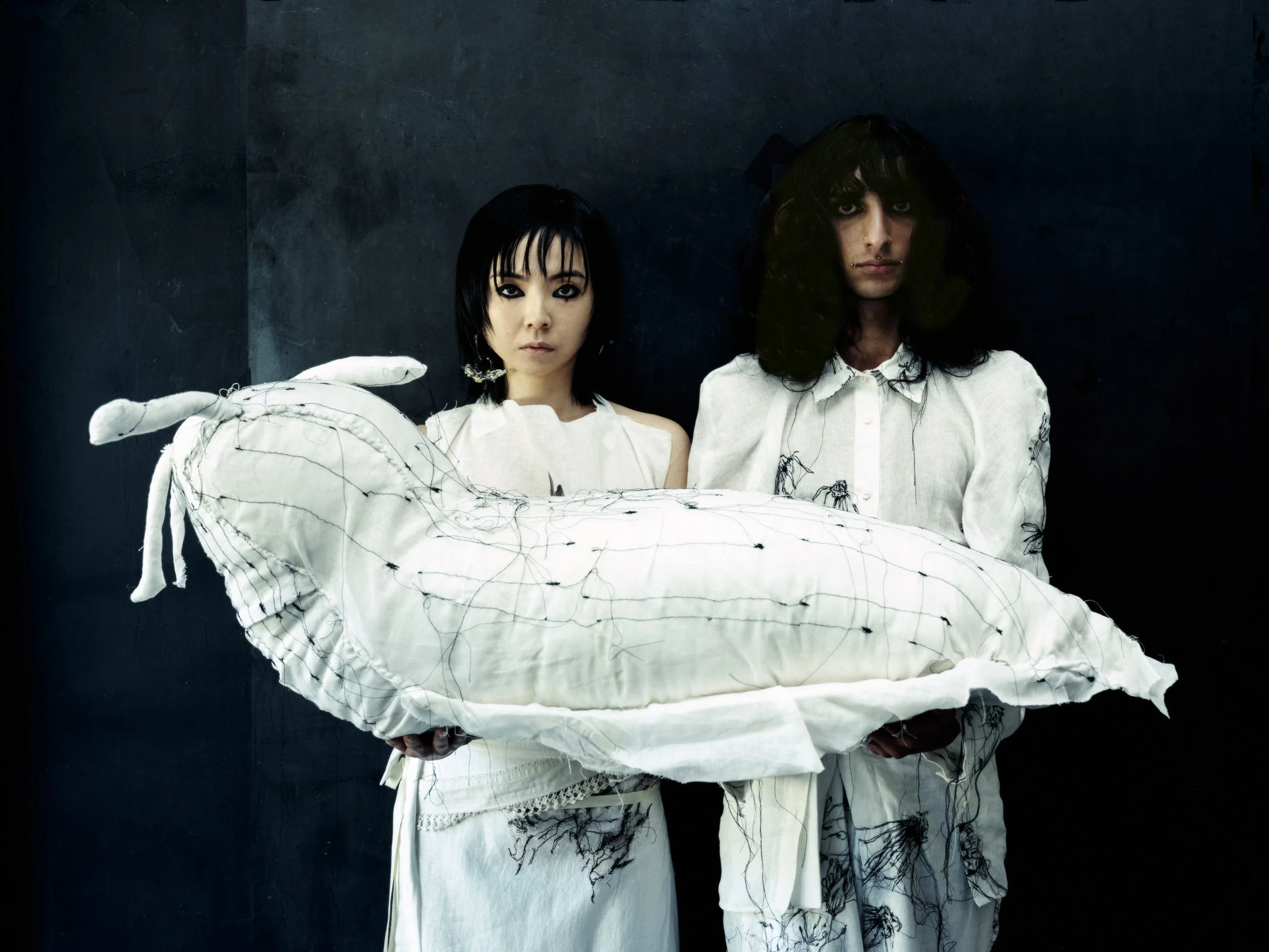 Zwei Personen halten ein humanoides Kuscheltier in Form eines Siddhartha-Guitars, die eine schwarze Haarpracht und weiße Kleidung tragen, vor einem dunklen Hintergrund.